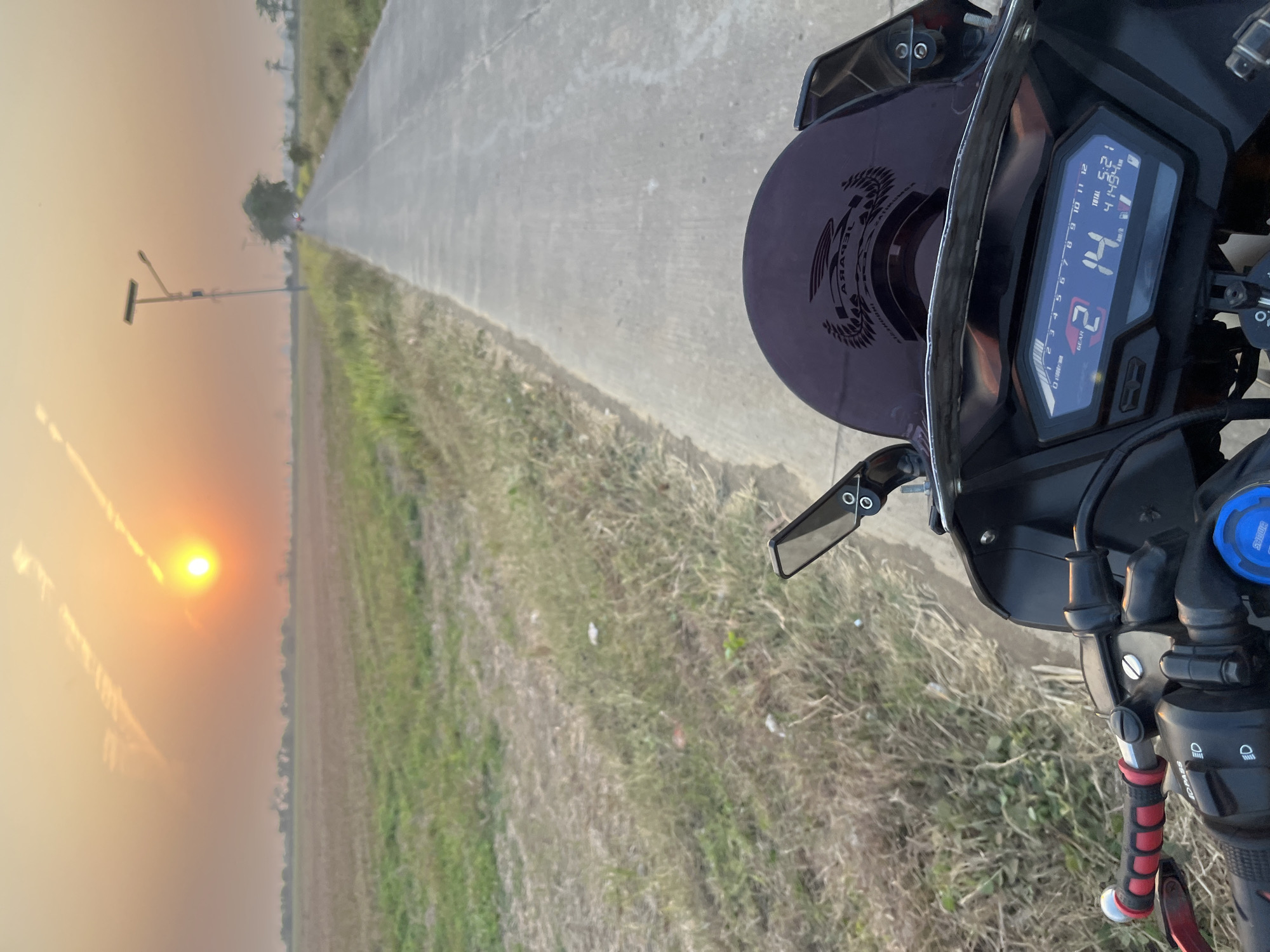 The image depicts a first-person perspective from a motorcycle in Troso, Indonesia, at sunset. A paved road stretches into the distance, flanked by dry grass and cultivated fields. The sun, low on the horizon, casts a warm, orange glow across the sky, with faint contrails visible. A lamppost with a solar panel stands on the right side of the road. The motorcycle's dashboard is visible in the foreground, showing the gear is "2", the speed is "14" km/h, the total time is "5:21", and the odometer reads "41494". The handlebars and a portion of the windshield are also in view. The overall scene suggests a peaceful, rural setting at dusk.