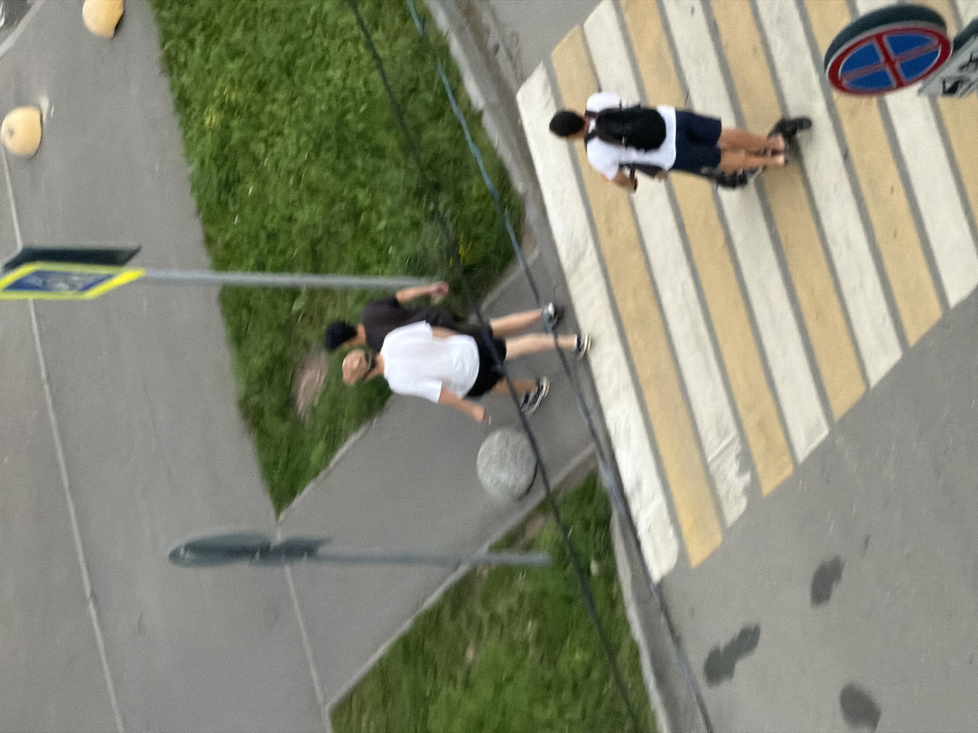 An aerial view captures a street scene in Kazan, Russia, featuring a pedestrian crossing, a sidewalk, and a grassy area. On the sidewalk, two individuals are walking; one wears a white t-shirt and dark shorts, while the other is dressed in a dark t-shirt and dark shorts. A person on an electric scooter, wearing a white t-shirt and dark shorts, is crossing the pedestrian zone. Visible on the side of the road are traffic bollards and a pedestrian crossing sign. The ground is marked with tire tracks and smudges.
