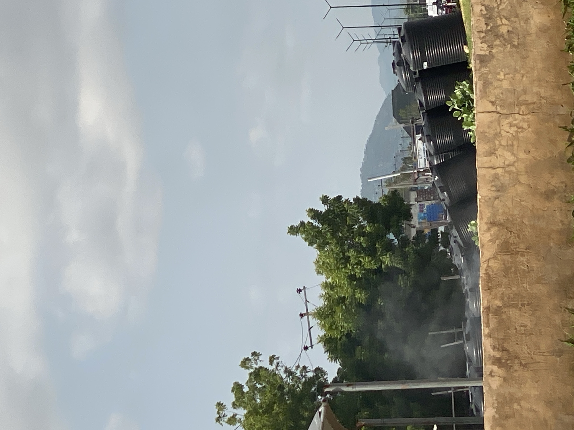 The image depicts an outdoor scene in Jalingo, Nigeria, with a focus on a collection of large, black plastic water tanks. These tanks are arranged in a row on a slightly sloped surface, with some appearing to be new and others possibly in use. A low, textured concrete wall runs along the bottom of the frame, separating the foreground from the area where the tanks are located. Behind the tanks, lush green trees and foliage are visible, suggesting a natural or semi-natural environment. In the background, a gentle mountain range rises against a sky filled with scattered clouds. Several streetlights with a distinctive design are also present in the middle ground, indicating an urban or semi-urban setting. A small amount of smoke or steam is visible in the left-central portion of the image, near some wooden structures and the trees, the source of which is unclear. The lighting suggests it is daytime.