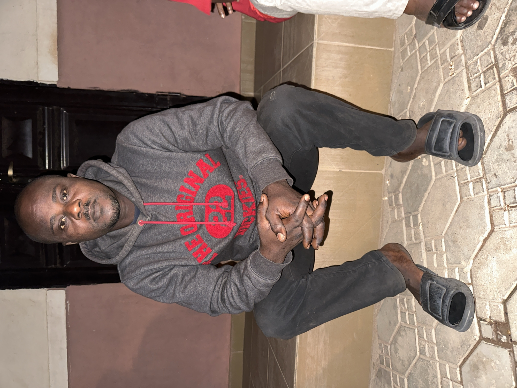 A full shot shows a Black man in a gray hooded sweatshirt with red lettering and black pants, sitting on a concrete step with a tiled walkway in front. The man's hands are clasped in his lap, and he is looking directly at the camera. He is wearing black slide sandals. The background appears to be a building facade with a dark wooden door. The lighting suggests it is evening or nighttime. The location is described as Narayi, Nigeria.