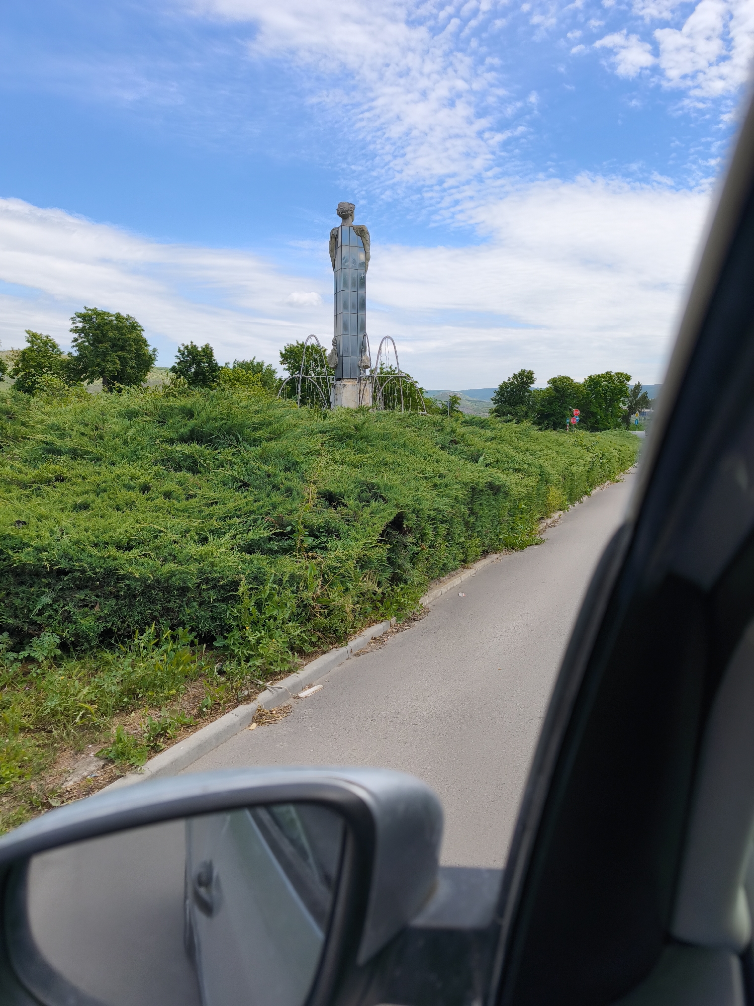 The image shows a view from inside a car, partially framing the car door and side mirror. Outside, a narrow asphalt road runs parallel to a large, dense hedge of green bushes. Beyond the hedge, there is a prominent silver statue of a human figure atop a tall, rectangular base, possibly made of glass or polished stone. A metal archway or fountain structure surrounds the statue's base. The background includes scattered trees, a slightly hilly landscape, and a blue sky with scattered clouds. The location appears to be the city of Unknown, Unknown. A "STOP" sign is visible in the background.