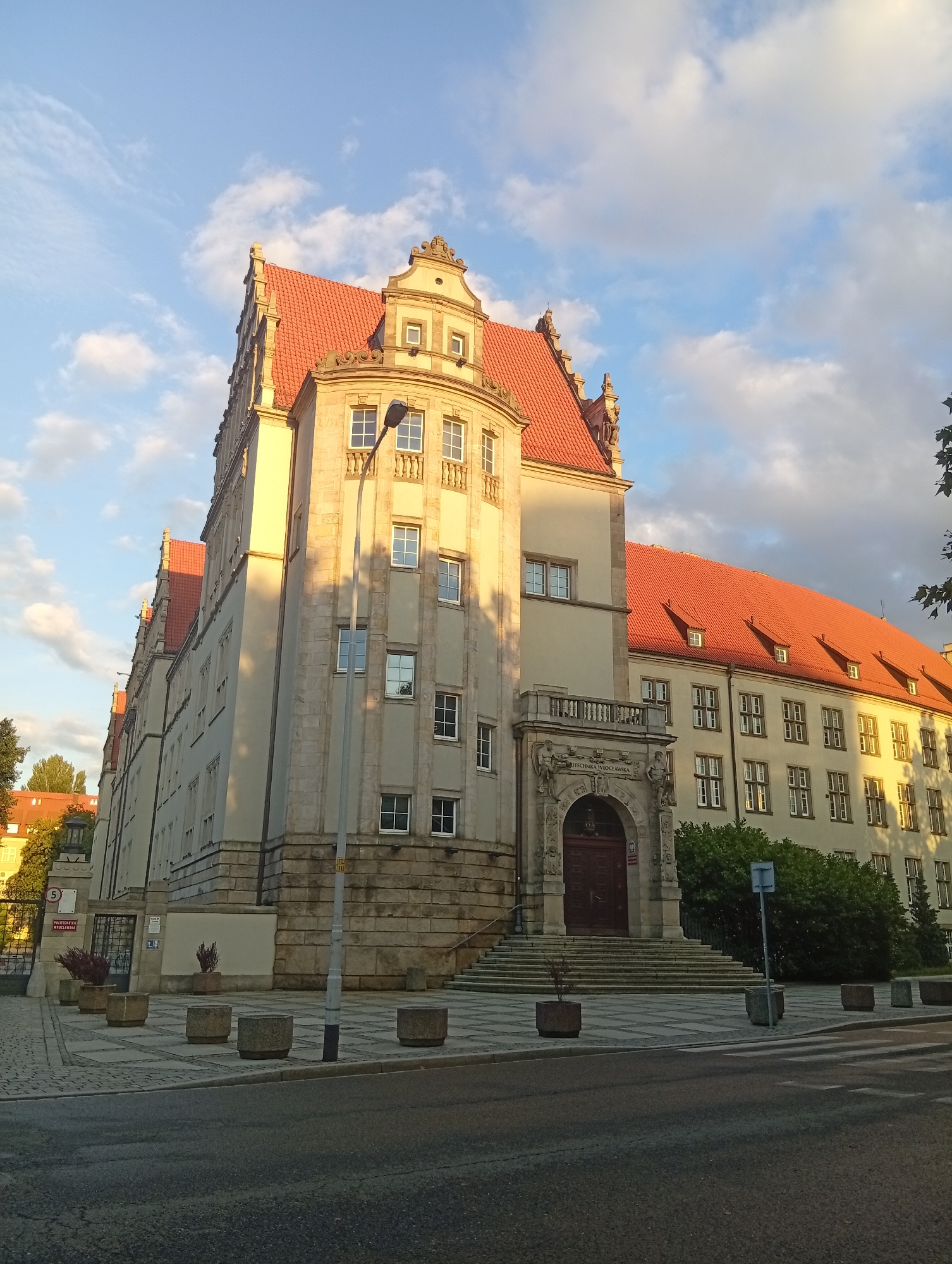 The image shows a sunlit, ornate building with red tile roofs, located in Wrocław, Poland. The building, identified as the Politechnika Wrocławska (Wrocław University of Science and Technology), is characterized by its multi-story facade, numerous windows, and a grand entrance with stone carvings. In the foreground, a street and sidewalk are visible, with concrete planters and a pedestrian crossing. The sky above is blue with scattered clouds, and the setting sun casts long shadows across the building and the pavement. The architecture suggests a historical or institutional structure, possibly a university campus building.