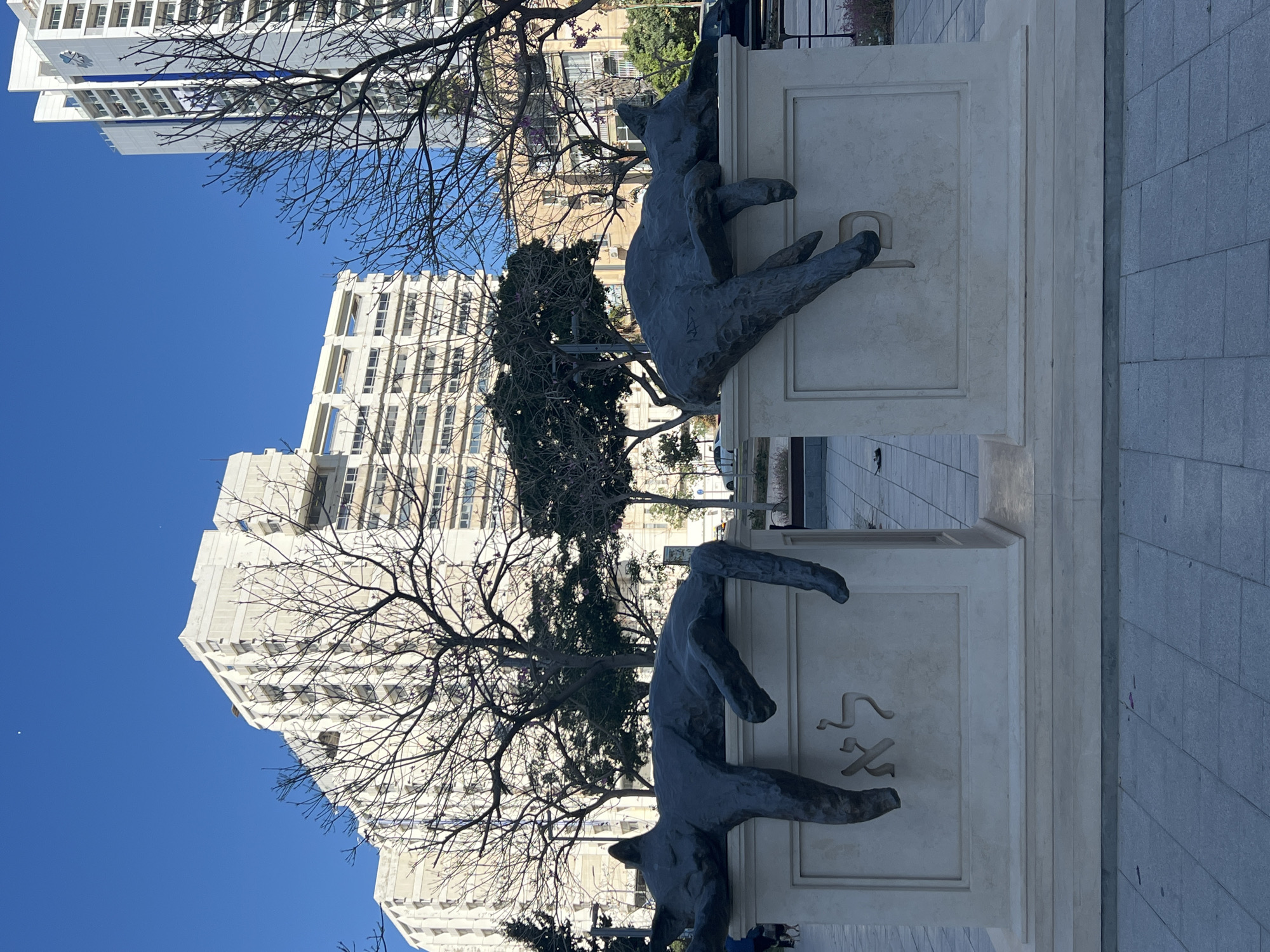 The image shows a sculpture of two dogs climbing over a wall. The wall is inscribed with Hebrew letters. In the background, there are tall buildings and trees, and the sky is blue. The location is likely in Tel Aviv, Israel, as the style of the buildings and the Hebrew letters are characteristic of the city.
