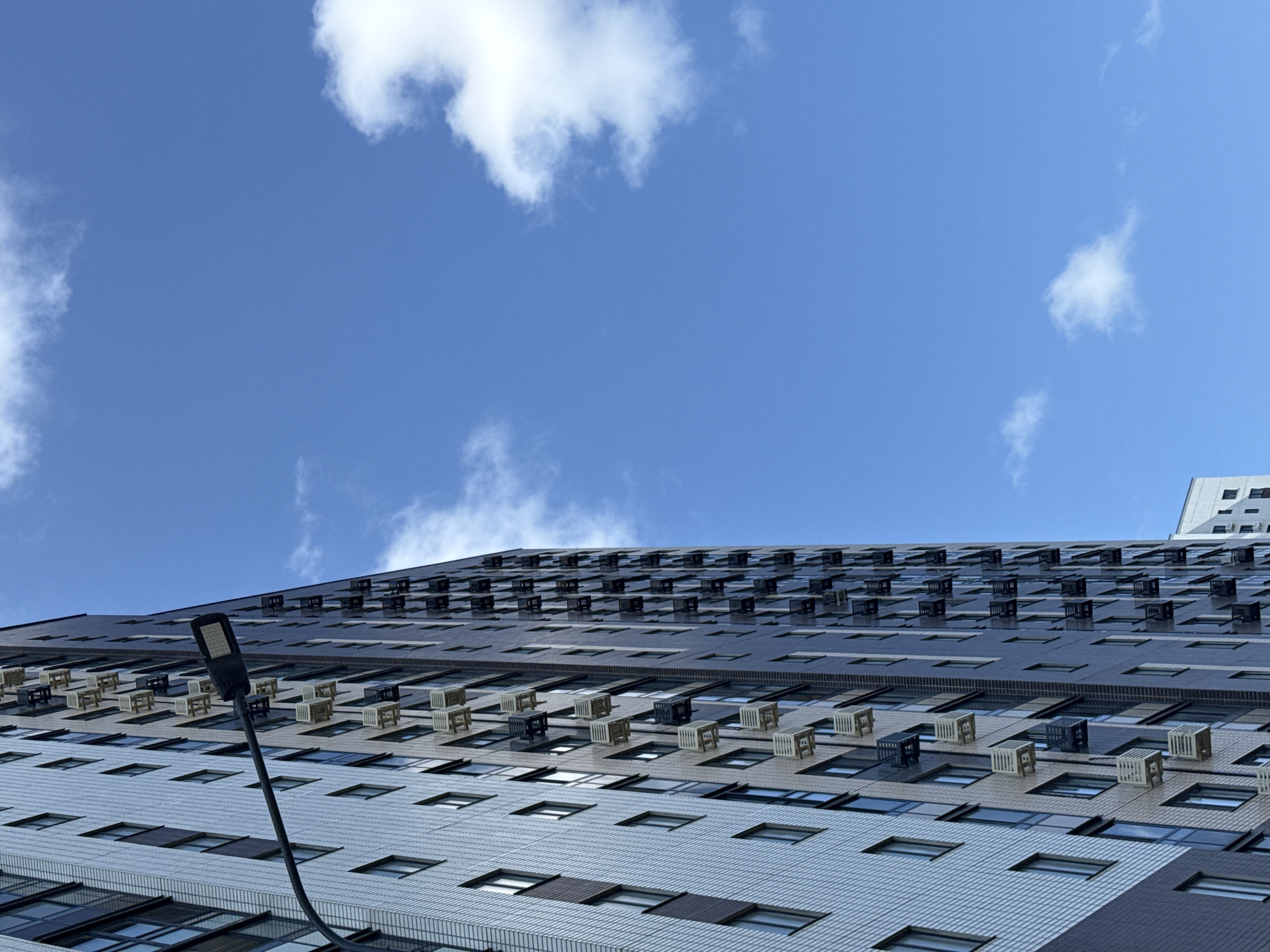 This is a medium shot, angled upward, of a tall, modern apartment building in Lyubertsy, Russia. The building's facade is a combination of light-colored textured tiles and darker, smooth panels. Numerous air conditioning units are mounted on the exterior walls, arranged in a regular pattern. A black street lamp with an LED fixture extends from the left side of the frame, arching over the building. The sky is a vibrant blue with scattered white, fluffy clouds. The image captures an urban architectural scene under daylight conditions.