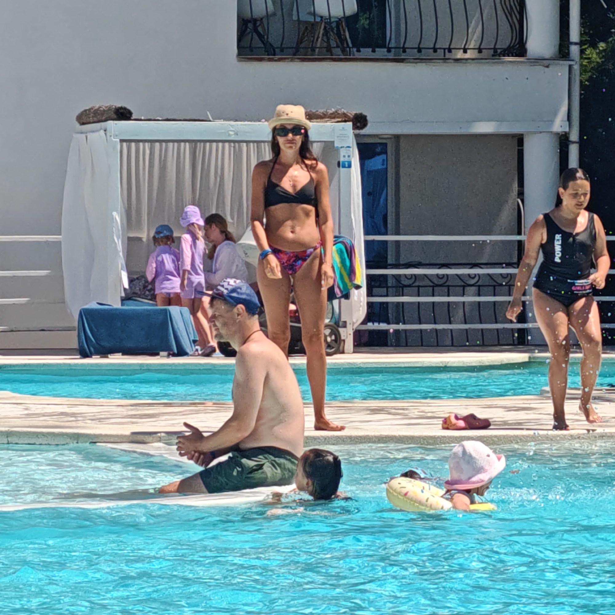 The image depicts a sunny day at a swimming pool. Several people are present, including adults and children. In the foreground, a man wearing a blue and white checkered cap and green swim shorts is partially submerged in the pool, facing left. He appears to be talking or interacting with someone out of frame. To his right, a young child wearing a pink hat and a yellow float with a donut pattern is also in the pool, facing away from the camera.
In the middle ground, a woman in a black bikini and a straw hat is standing on the pool deck, facing the camera. She is wearing sunglasses and a necklace. To her left, a group of children in purple swimsuits are standing near a blue-covered table.
In the background, another young woman in a black swimsuit with the word "POWER" visible on the strap is walking on the pool deck. She has her hair pulled back and is looking to her left. A white building with railings and some greenery is visible behind the people. The water in the pool is bright blue and appears to be calm, with some ripples. The overall impression is a relaxed and enjoyable day at the pool.