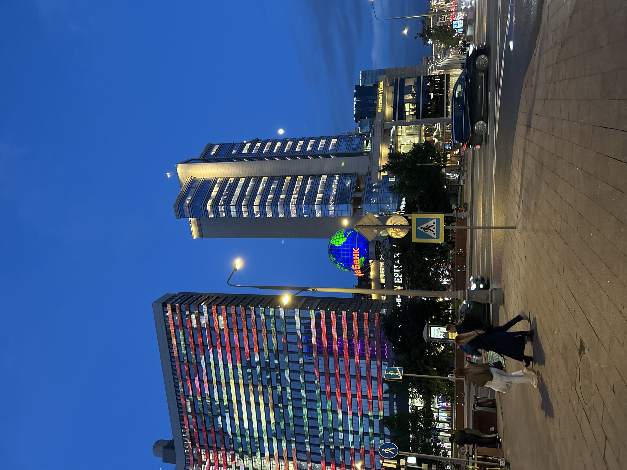The image depicts a nighttime scene in a city, likely taken from a street-level perspective. The scene is characterized by modern architecture and urban elements.
There are several buildings in the shot. One appears to be a skyscraper with illuminated windows, suggesting office spaces. Another building is notable for its exterior being lit up with a grid pattern of red, white, blue, and green lights. A third building has a sign that reads "РЕСТОРАН YUMA" (RESTAURANT YUMA).
Streetlights are present, casting a warm glow on the surroundings. There is also a large globe-shaped structure with the word "абанк" (ABANK) displayed on it, possibly advertising a bank.
Several people are visible on the sidewalk, appearing to be pedestrians walking or standing. A car is on the road. There are also pedestrian crossing signs visible.