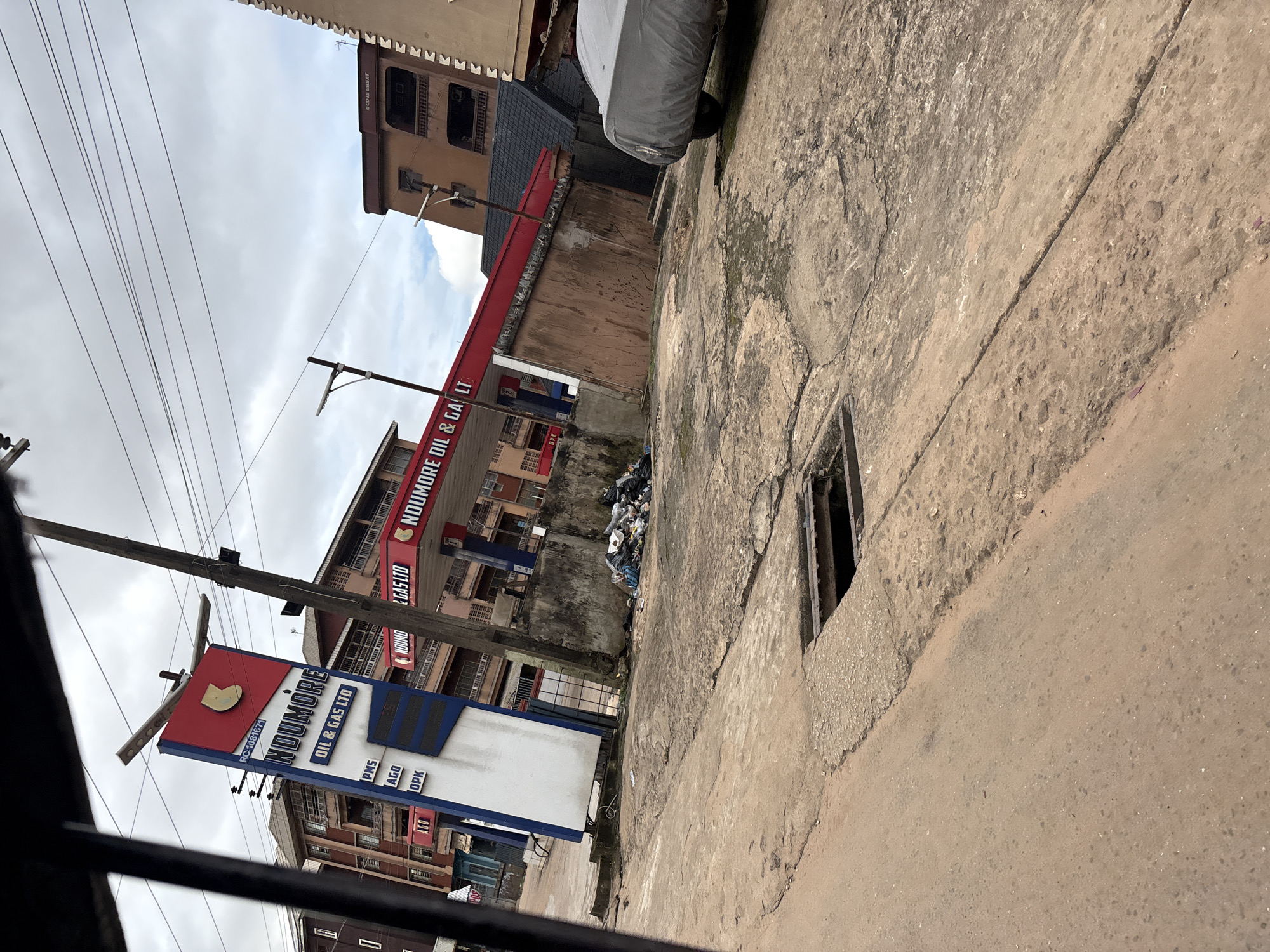 The image depicts an outdoor scene at a NouMore Oil & Gas Ltd. station in Nkpor, Nigeria. A tall, red and blue sign with the station's name and "OIL & GAS LTD" is prominently displayed. Below the name, prices for PMS, AGO, and DPK are listed. A red canopy with the same station branding extends over a concrete area, where a car covered with a gray tarp is parked on the right side. A pile of refuse bags is accumulated near a concrete wall on the left. The ground is paved with concrete, showing signs of wear and tear, with a visible open drain cover in the foreground. Buildings, some with multiple stories, are visible in the background, and a utility pole with overhead wires stands near the gas station sign. The sky is overcast, suggesting a cloudy day.