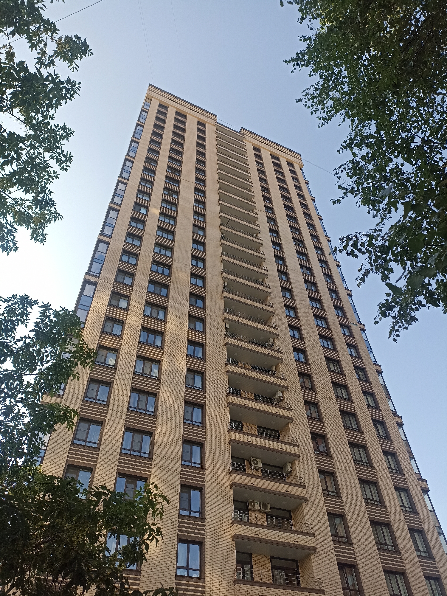 This is a low-angle, full shot of a tall, modern apartment building in Novosibirsk, Russia, on a clear day. The building, with a light beige brick facade and dark brown trim around the windows, dominates the frame, stretching towards a pale blue sky. Rows of windows, appearing uniformly dark, are visible on each level. Balconies with dark railings and light-colored floors extend from many of the upper floors. Several outdoor air conditioning units are mounted on some of these balconies. The building's structure is somewhat angled, giving it a dynamic appearance. On the left and right sides of the image, the tops of lush green trees with dark leaves are seen, partially framing the building against the sky. A thin, dark wire stretches across the upper portion of the sky, possibly a power line or antenna wire. No people are visible in the image. The scene depicts a typical urban residential building in a city environment.