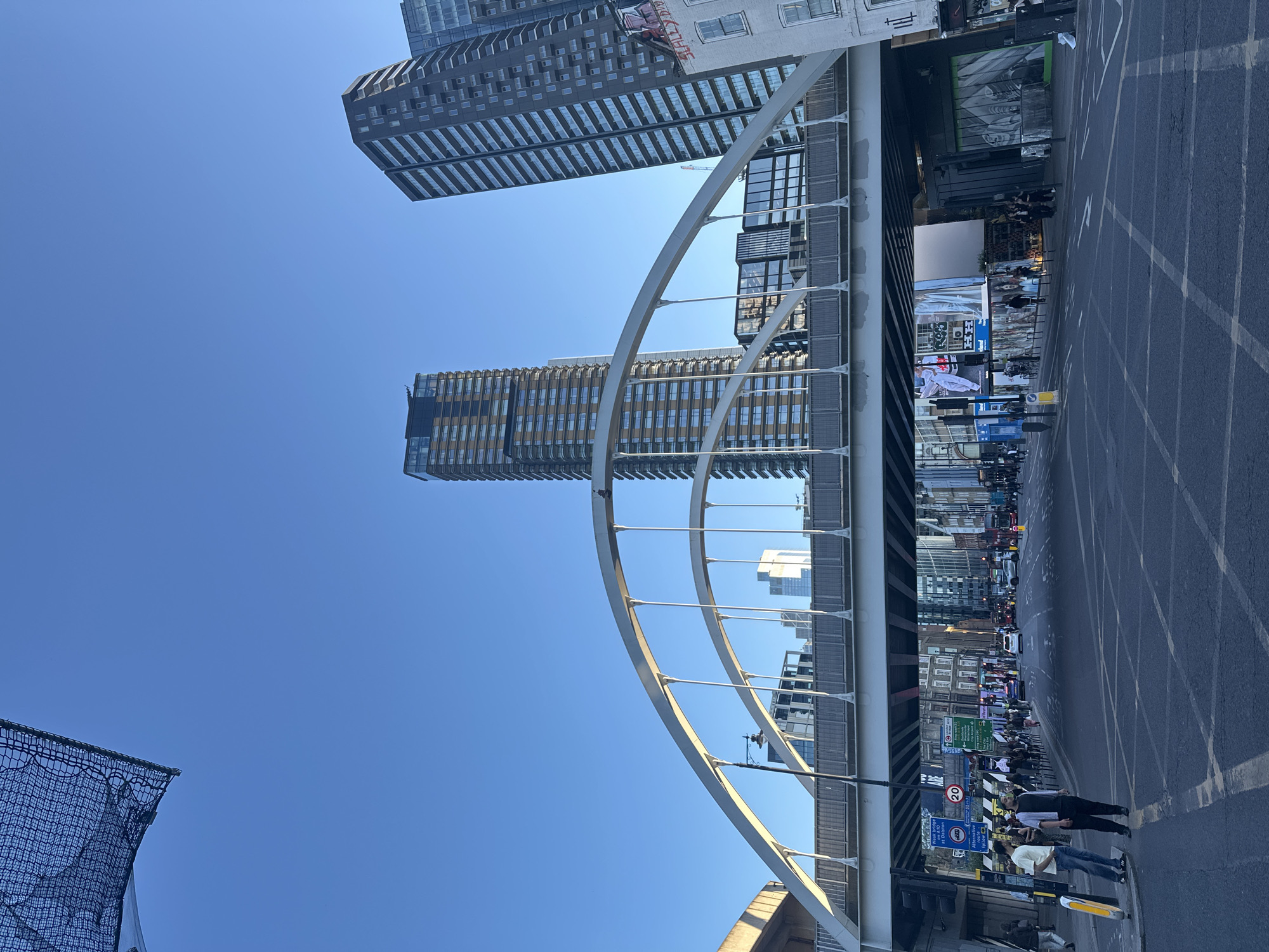 The image depicts a street scene in London, United Kingdom, under a clear blue sky. A modern, white, arched pedestrian bridge dominates the foreground, framing views of two tall skyscraper buildings. The buildings have a modern design. Below the bridge, there is a busy street with cars and pedestrians. Signage, including traffic and directional signs, is visible. People are walking on the sidewalk. The street has markings, suggesting a designated route for vehicles. A net is on the bottom left of the image. The overall impression is that of a busy urban environment in a major city.