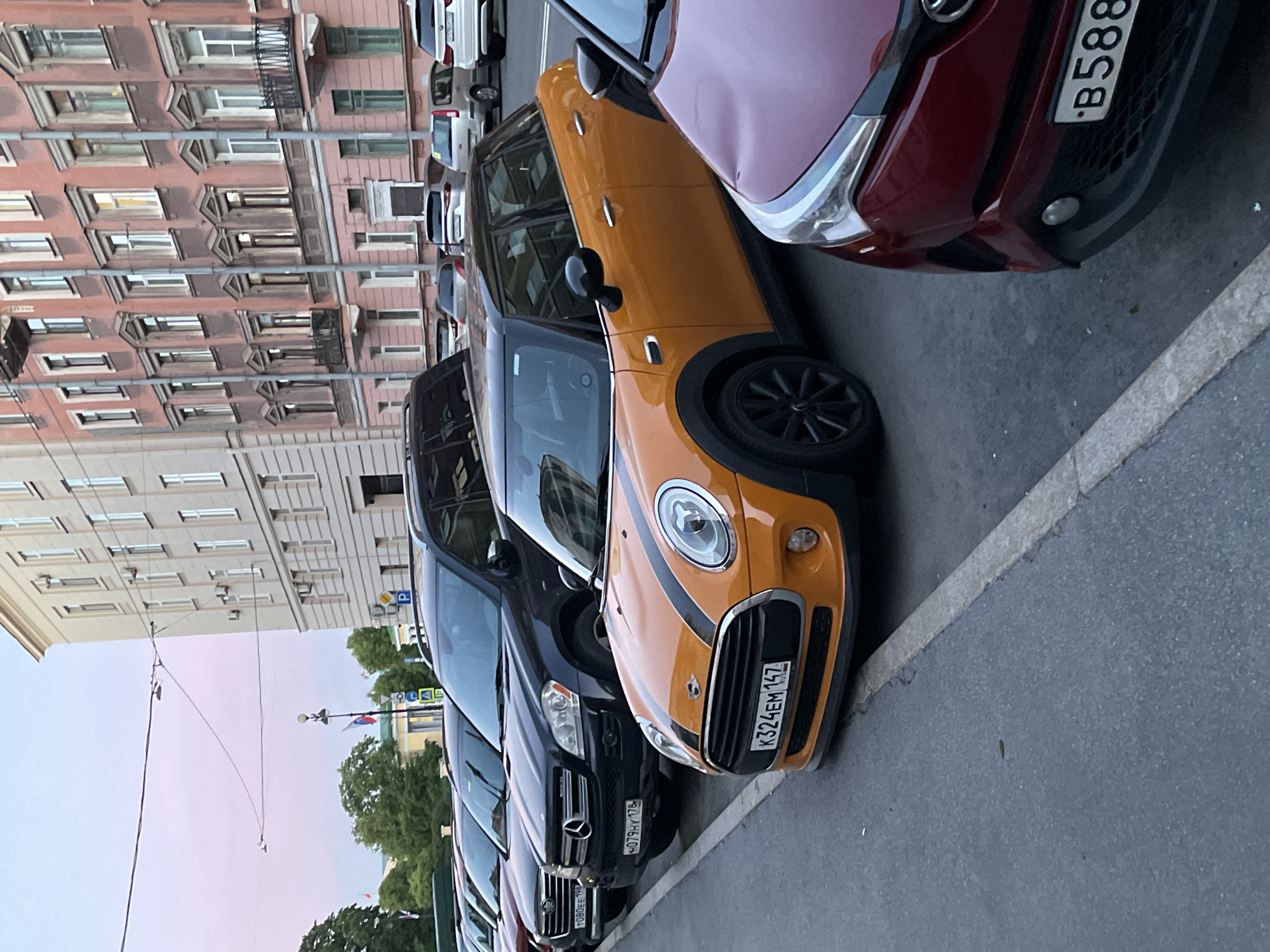 A line of parked cars is visible along a city street. In the foreground, a red car is partially visible, with its front bumper and headlights showing. Next to it, an orange Mini Cooper with black racing stripes is parked, displaying a Russian license plate with the characters "K324EM 147 RUS". Behind the Mini Cooper, a black Mercedes-Benz SUV is parked, also with a Russian license plate that reads "H079HY 178". Further down the street, other cars are parked, including white SUVs and sedans. The cars are parked parallel to a sidewalk. In the background, a multi-story building with ornate architecture and numerous windows is visible. The sky is a pale pink and purple, suggesting it is either sunrise or sunset. The architecture of the building and the presence of Russian license plates strongly indicate the location is Saint Petersburg, Russia. The scene depicts a typical urban streetscape with vehicles parked along the roadside. There are no people visible in the image.
