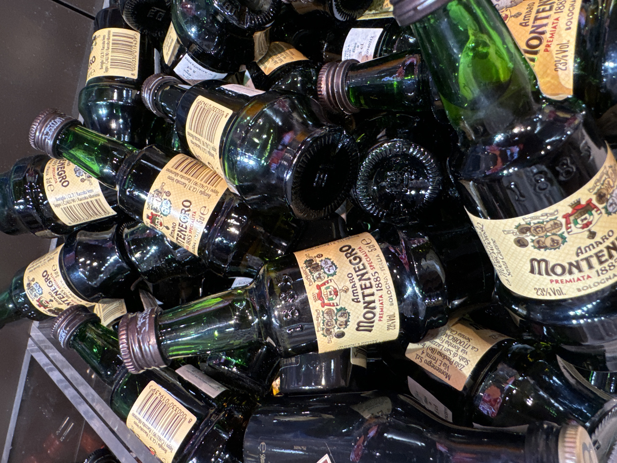 The image shows a pile of miniature bottles of Amaro Montenegro liqueur. The bottles are dark green glass with labels indicating the brand name, alcohol content (23% Vol.), and place of origin (Bologna, Italy). Some labels also include recycling information for the glass bottle and aluminum cap. The bottles are stacked haphazardly, filling the frame, and appear to be contained within a dark-colored bin or container, suggesting they may be for sale or in storage. The scene is indoors, under artificial lighting.