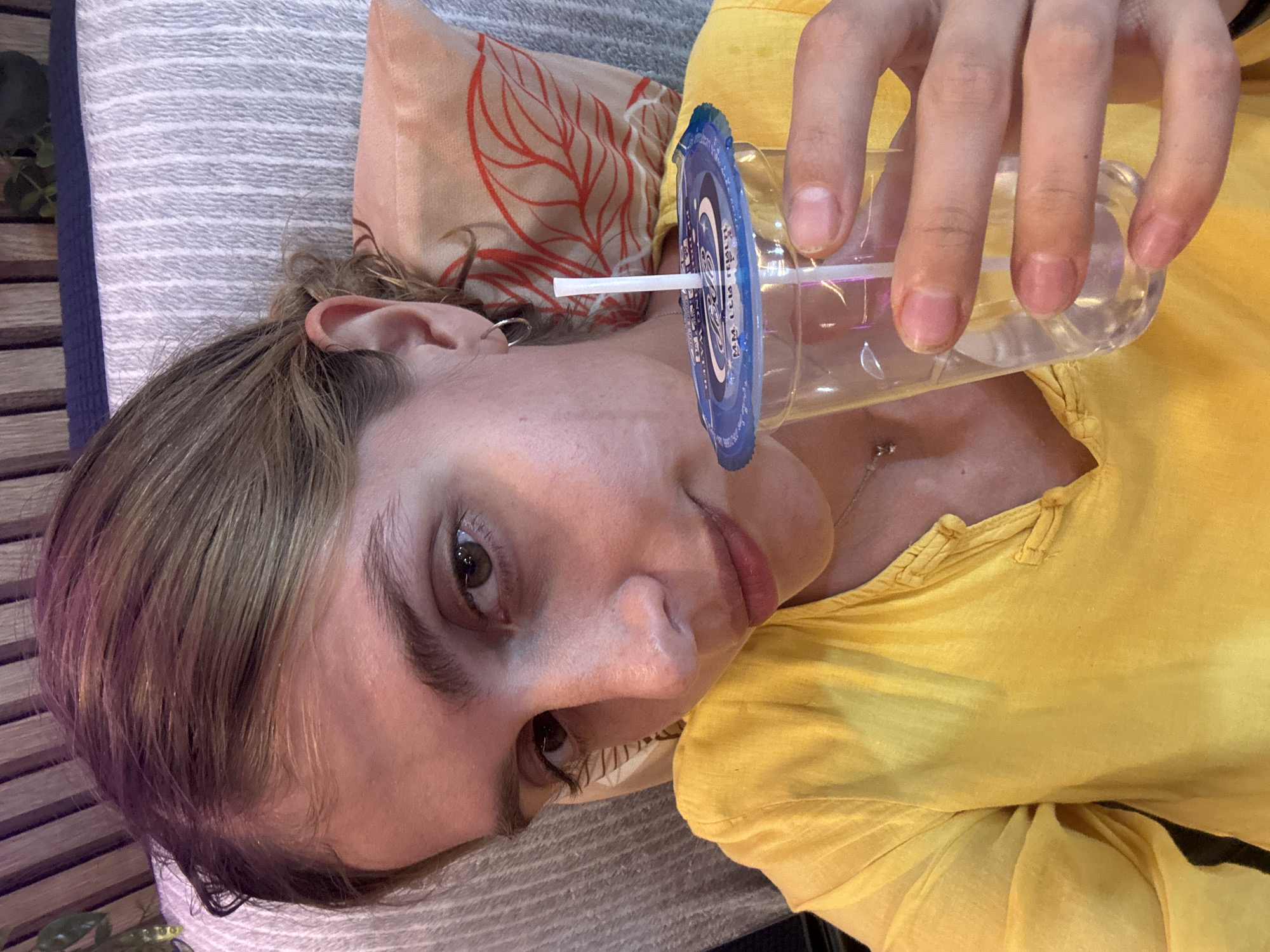 The image shows a person lying down, holding a clear plastic cup with a blue lid and a white straw. The cup appears to contain water. The person is wearing a yellow shirt. The person is resting on a striped cushion, with a patterned pillow nearby. The image provides no information about the location or city.
