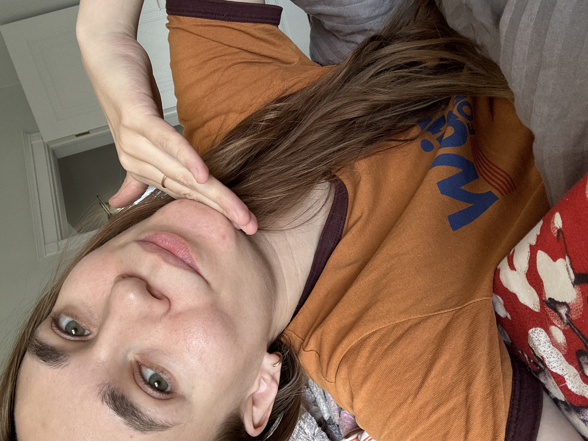A woman is lying down, facing the camera at an angle. She is wearing an orange t-shirt with brown trim and a blue and red logo. Her hair is brown, and she has green eyes. Her left hand is touching her face. She is wearing a gold ring on her finger and a gold earring in her ear. The background shows a white door and what appears to be bedding and pillows. The location is not apparent.