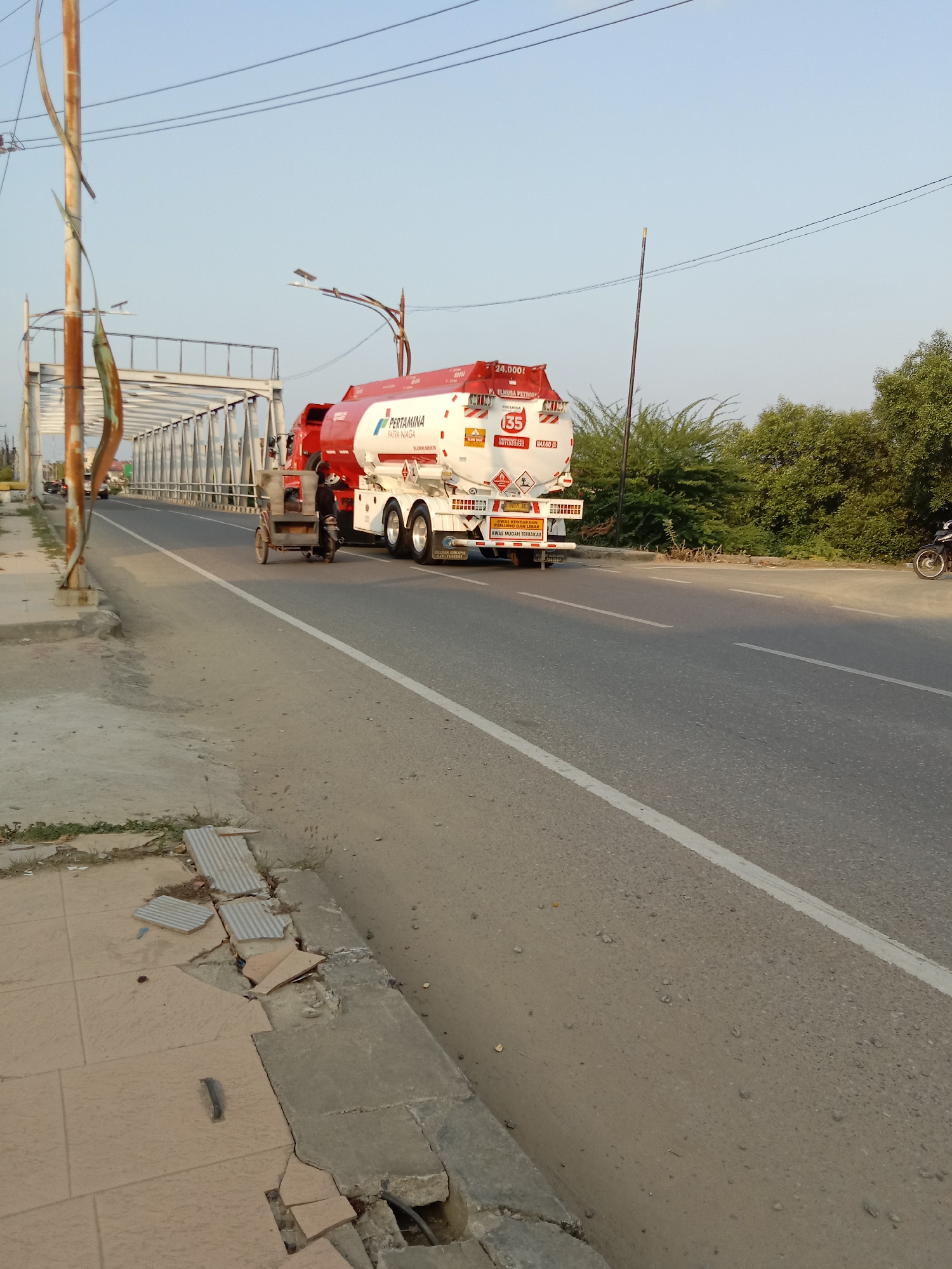 This is a full-body shot of a large white and red Pertamina fuel tanker truck driving on a paved road over a bridge in Lhokseumawe, Indonesia. The truck has the Pertamina logo on its side, along with other text and safety symbols. In front of the truck, a person is riding a three-wheeled motorcycle with a cargo box. The road is asphalt and has a white dividing line. To the left of the road is a sidewalk with broken tiles and some vegetation. To the right of the road, there are green trees and foliage. The sky is clear and blue. The scene appears to be captured during the day, likely late afternoon due to the warm lighting.