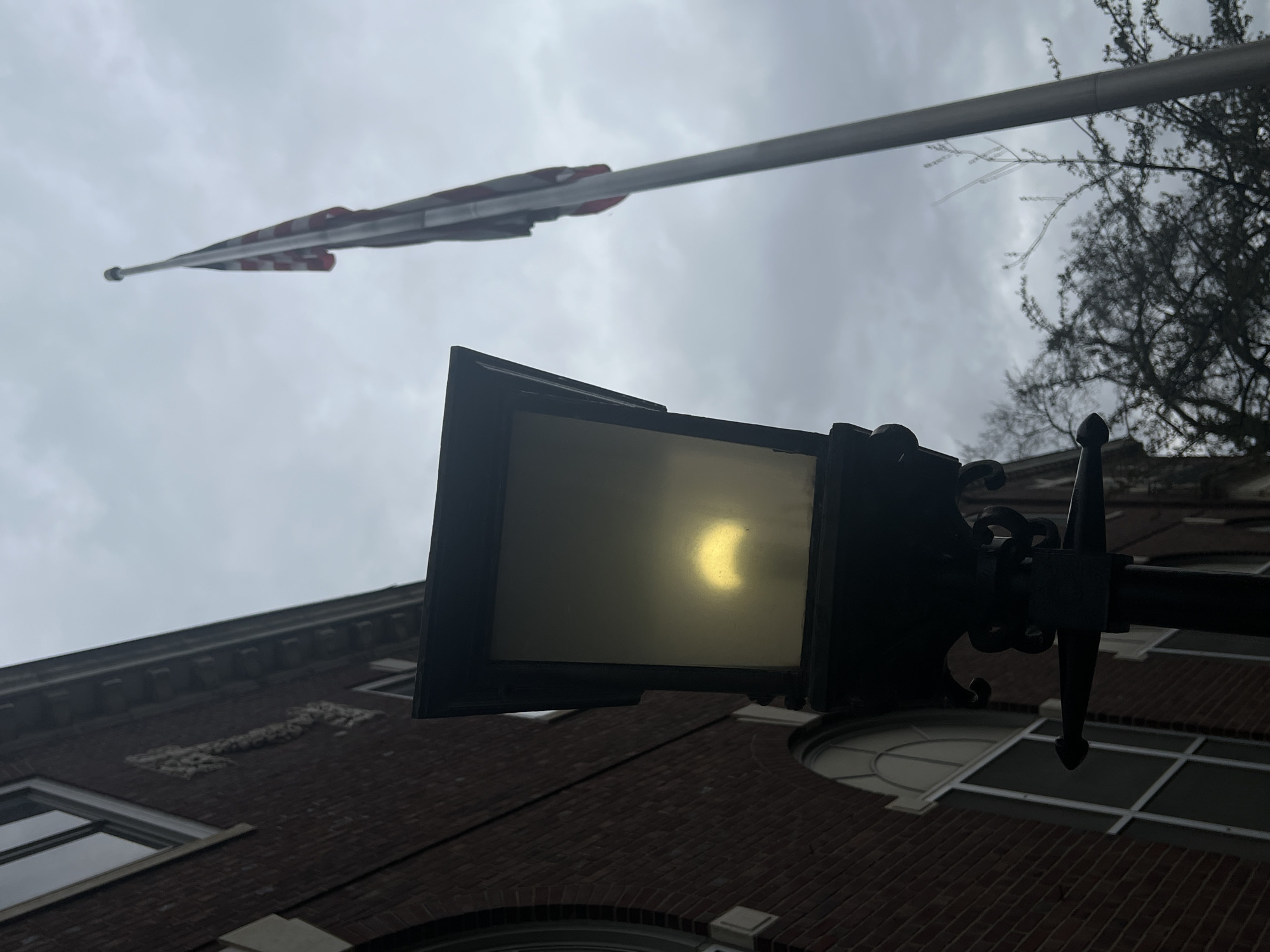 The photograph, taken in the City of Newburgh, United States of America, appears to capture a solar eclipse through the lens of a street lamp's light. The lamp is centrally positioned, casting an eerie glow with the partial eclipse visible within the lamp's light. Above the lamp, a slightly out-of-focus American flag is seen waving atop a pole against a cloudy sky. To the right of the flag, tree branches are visible. In the lower section of the photo, a brick building with multiple windows can be observed.