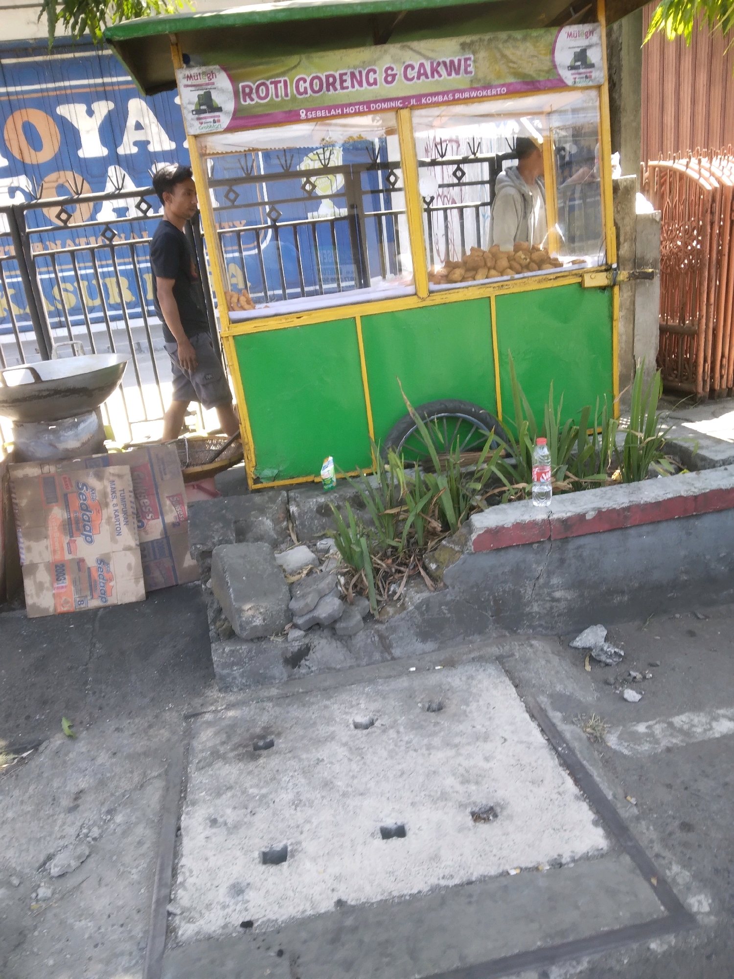 The image shows a street scene in Purwokerto, Indonesia. A food cart selling "Roti Goreng & Cakwe" (fried bread and fritters) is prominently displayed, with a sign indicating its location as "Jl. Hotel Dominic - Jl. Kombas Purwokerto." A young man in a black t-shirt and grey shorts is standing to the left of the cart, looking to the side. Another person, wearing a light-colored hooded jacket, is visible inside the cart, attending to the food. In front of the cart, there are stacks of cardboard boxes, one of which appears to be from the brand "Sedaap." A large wok-like cooking pan is placed on top of some of these boxes. The ground in front of the cart is paved with concrete, and there are some plants growing in a planter box. A plastic water bottle is visible near the plants. To the right of the cart, a corrugated metal fence or gate is present. The overall environment suggests an outdoor commercial area on a street.
