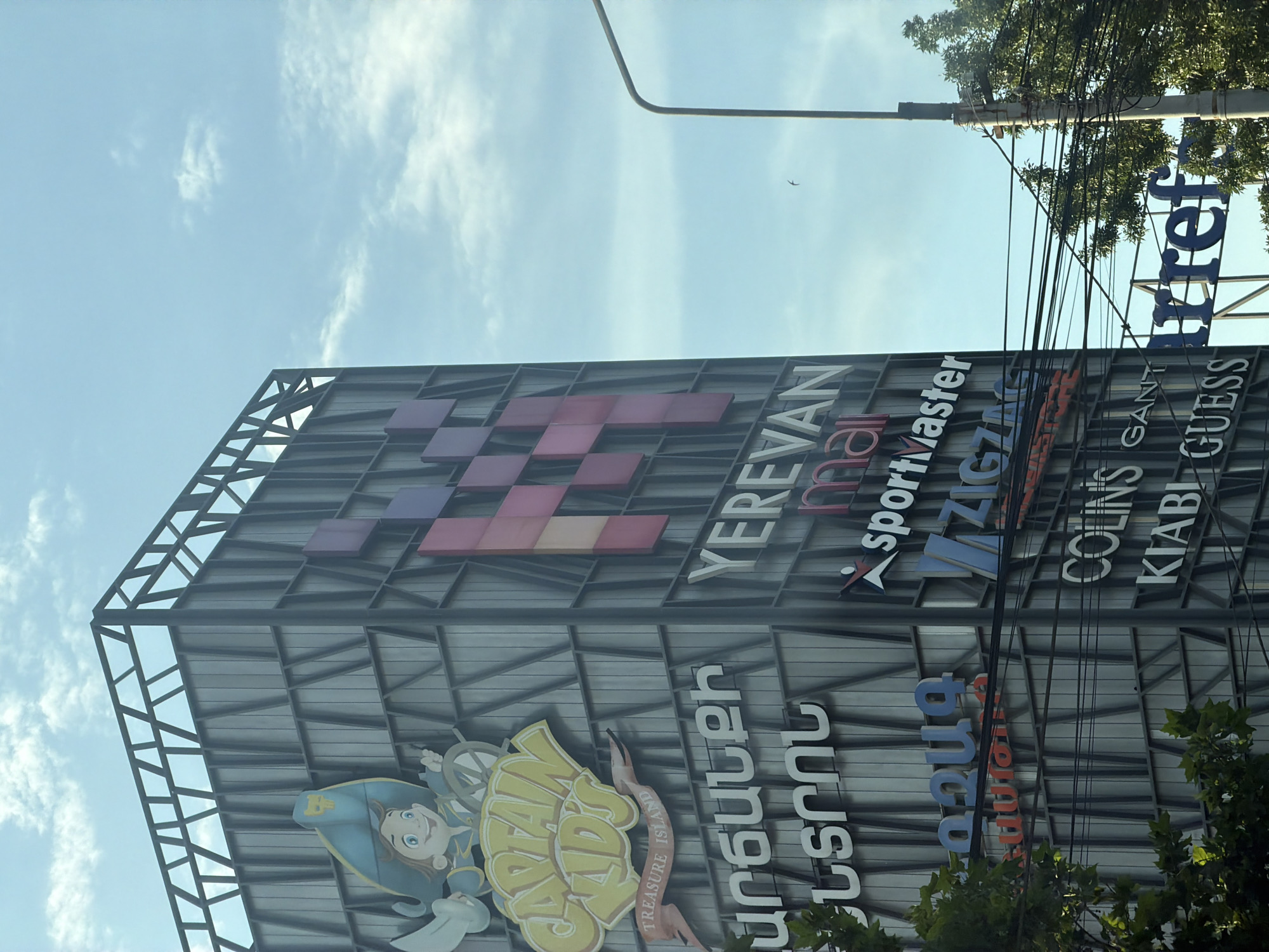 The image shows a close-up of a tall, modern building in Yerevan, Armenia, with multiple signs for various shops. The building is a light grey color with a geometric pattern of darker grey lines. On the left side of the building, there is a large, colorful sign for "Captain Kid's Treasure Island." Below this, there are signs in Armenian script, and then signs for "Yerevan mall," "sportmaster," "ZIGZAG," "COLIN'S," and "GUESS." To the far right, a sign for "Carrefour" is partially visible. The sky above is blue with scattered white clouds, and a lamppost is visible on the right side of the image, with power lines running across the frame. There are trees and greenery at the bottom of the image.