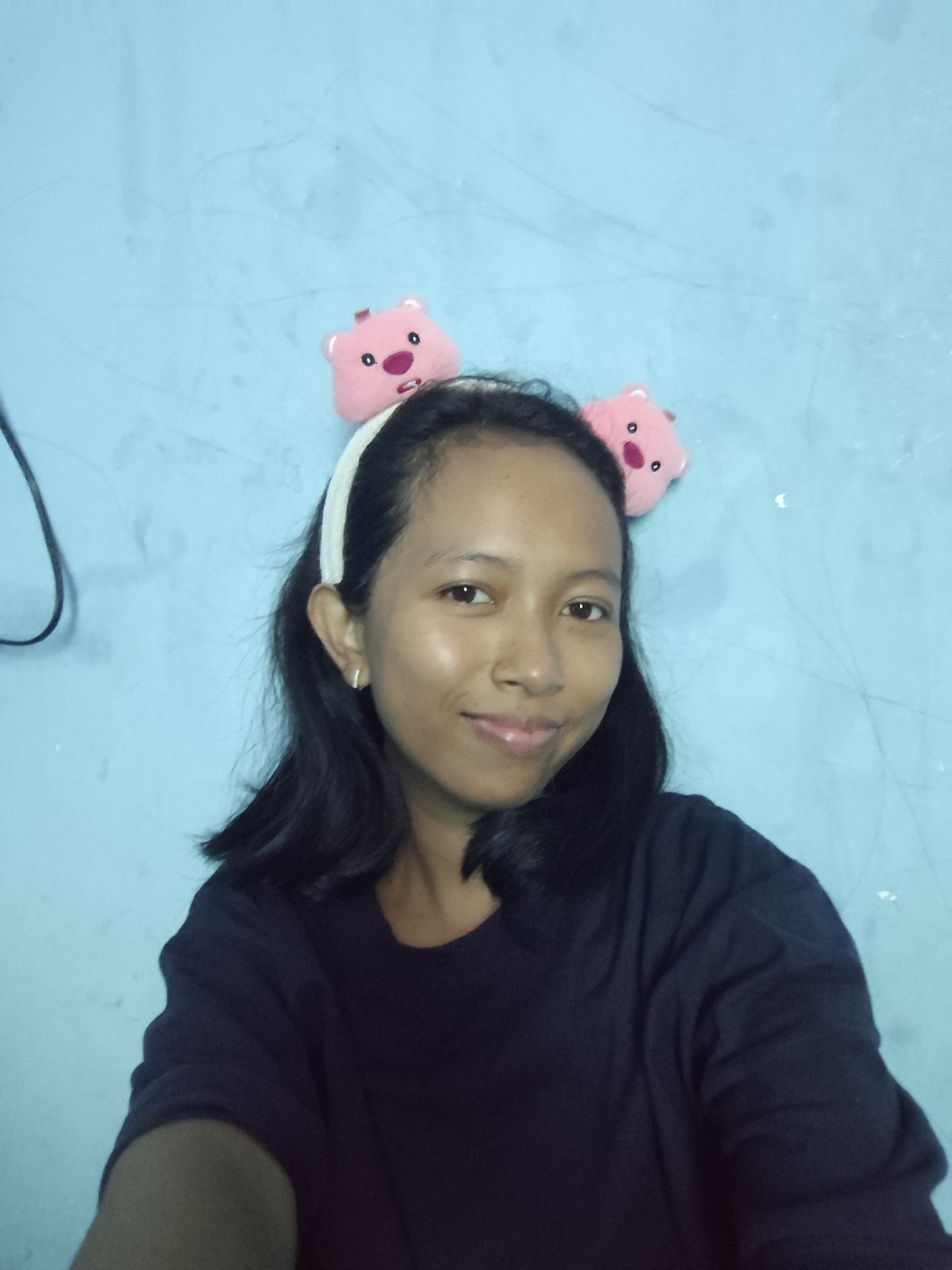 A young woman is captured from the chest up in a close-up, waist-up shot against a light blue, textured wall. She is wearing a dark blue long-sleeved shirt and a white headband adorned with two small, pink stuffed animal ears. The woman has shoulder-length, dark hair and is looking directly at the camera with a slight smile. Her skin appears to have a subtle sheen, possibly from natural light. A thin, dark wire is visible on the left side of the frame, extending from the upper left corner down to approximately her shoulder. The lighting is soft, and there are faint, horizontal lines and some speckling visible on the wall behind her. The overall atmosphere is casual and personal. The location is unknown.