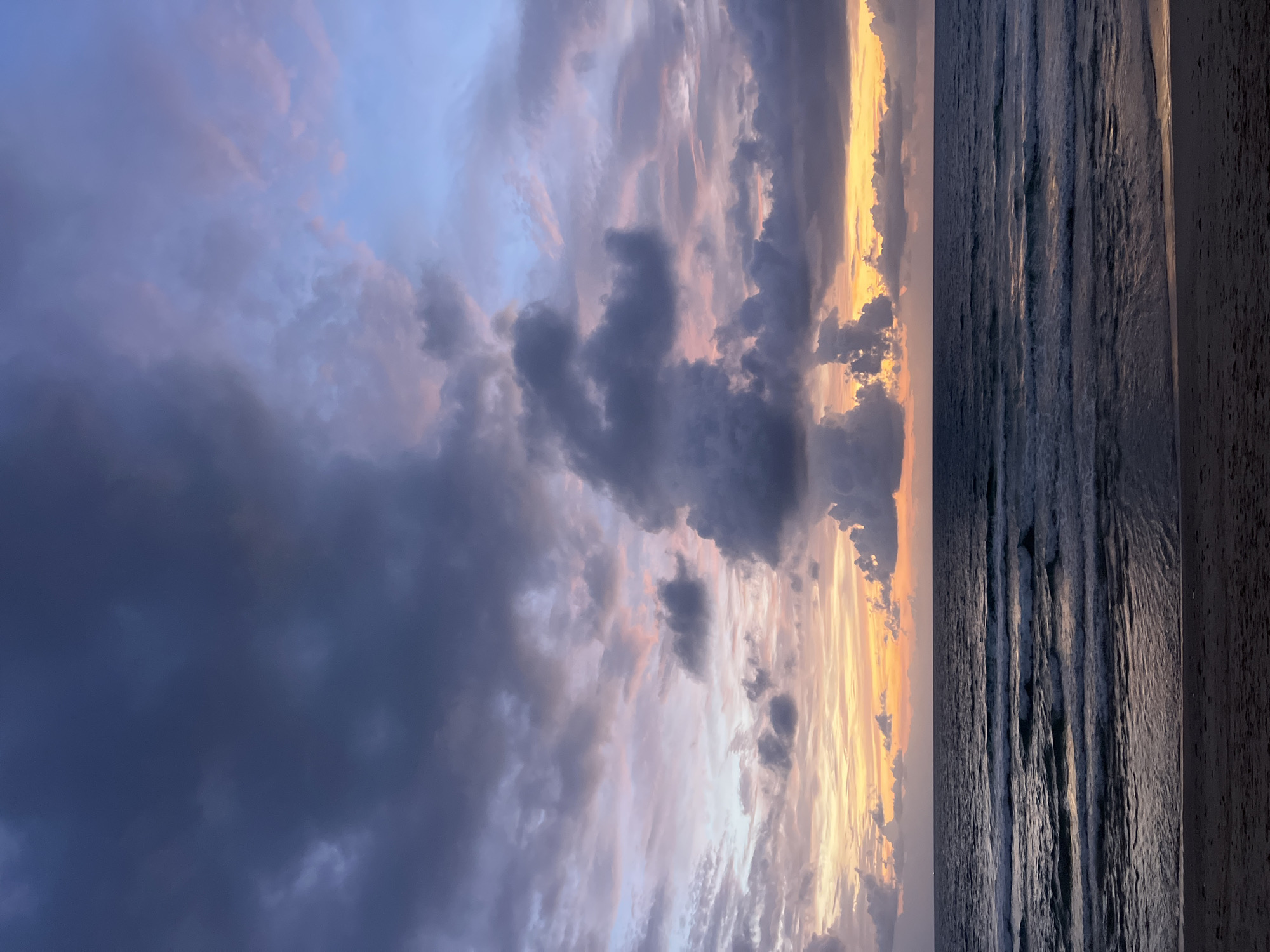 The image shows a beach scene in Choeng Thale, Thailand at twilight. The sky is filled with dramatic clouds in shades of gray, purple, pink, and yellow, indicating either sunrise or sunset. The ocean is dark and calm, with gentle waves lapping at the shore. The sandy beach is visible in the foreground, with subtle footprints. There are no people or man-made structures visible in the scene, focusing solely on the natural environment.