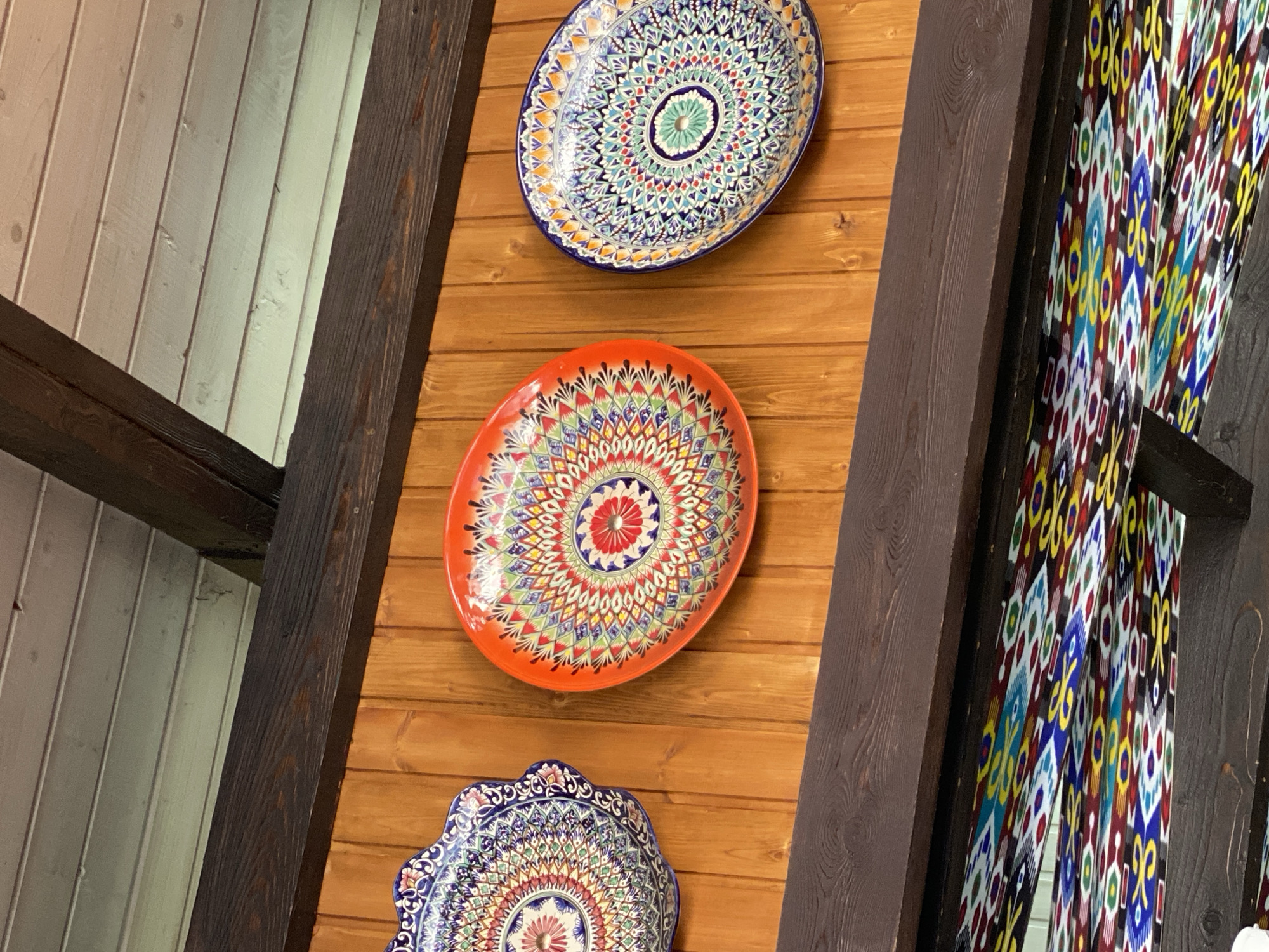 Three decorative ceramic plates with intricate, colorful patterns are displayed on a wooden wall. The plates are arranged from left to right, with the leftmost plate having a blue and white ornate edge. The middle plate is a vibrant orange with a radial mandala-like design in red, green, and yellow. The rightmost plate is oval-shaped with a blue border and a central green and teal design, surrounded by a pattern of multicolored geometric shapes. Above the plates, dark wooden beams and a light-colored paneled ceiling are visible. Below the plates, another dark wooden beam supports a ceiling adorned with a vibrant, patterned fabric or textile featuring geometric and abstract designs in blues, yellows, reds, and greens. The overall setting suggests an indoor decorative display, possibly in a cafe or shop.
