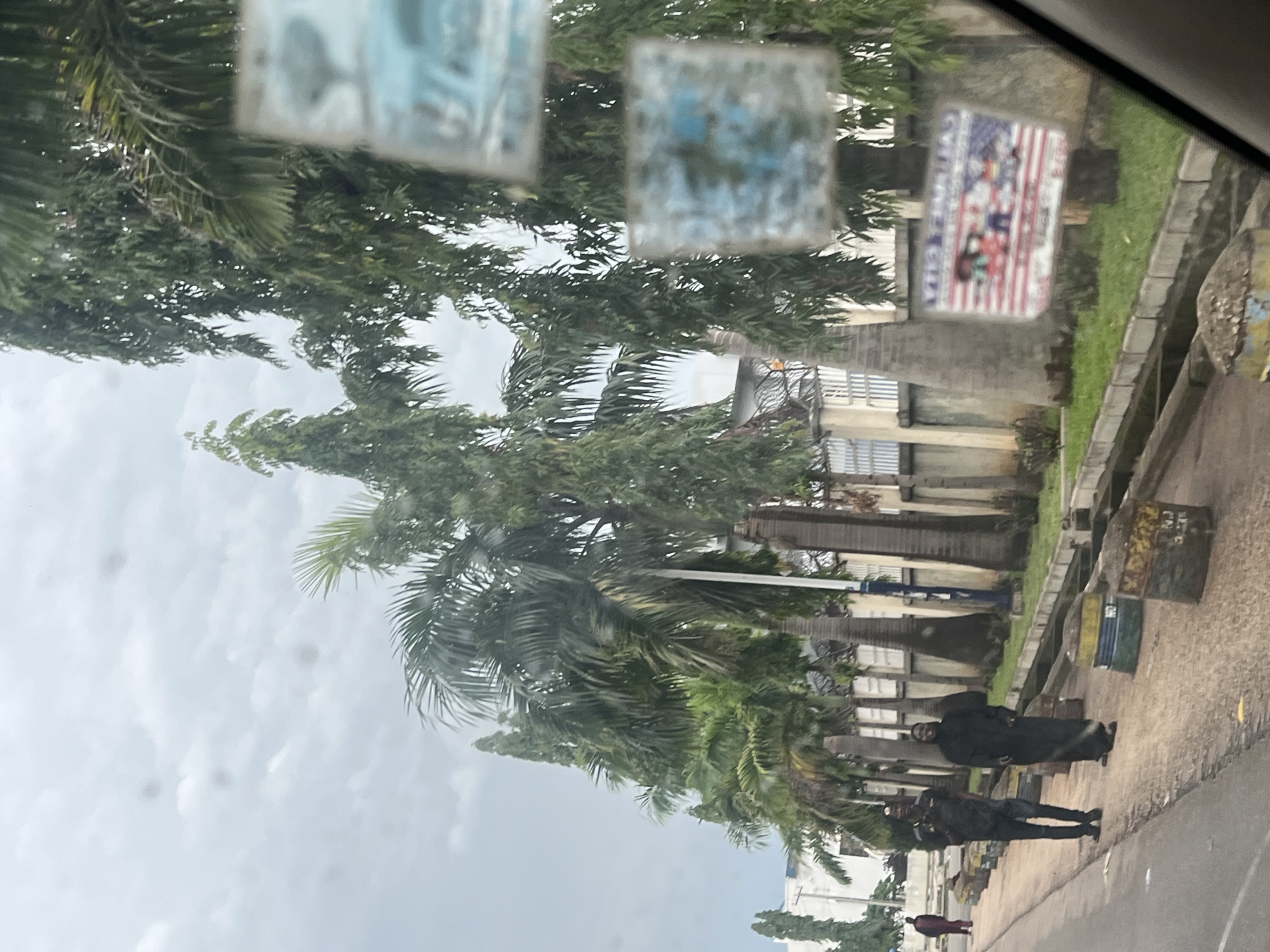 The image depicts a street scene in Jos, Nigeria, as viewed from inside a vehicle. The perspective is skewed, suggesting the photo was taken while moving or with a tilted camera. Lush green trees, including palm trees, dominate the foreground and midground, suggesting a tropical or subtropical environment. In the background, buildings with a mix of modern and older architectural styles are visible, bordered by a paved walkway and a roadside drain.
Several people are present on the walkway. One individual, dressed in dark clothing, stands near a building. Further down, two figures, also in dark attire, are positioned on the walkway. A fourth person in a maroon garment is seen further in the distance. The presence of these individuals and the roadside setting suggest an urban or semi-urban environment.
On the vehicle's windshield, two blurry rectangular stickers are visible. One of these stickers is more discernible, featuring a collage of images against a red and white striped background, resembling an American flag motif. It's difficult to make out specific details within the sticker due to its blurriness, but it appears to be a decorative or informational decal.
The sky is overcast with clouds, indicating a possibly cloudy or rainy day. The lighting is diffused, suggesting it is likely daytime, possibly morning or afternoon, but not bright sunlight. No specific activities or events are clearly depicted, beyond people simply standing or walking along the street. The overall impression is of a typical day on a street in Jos.