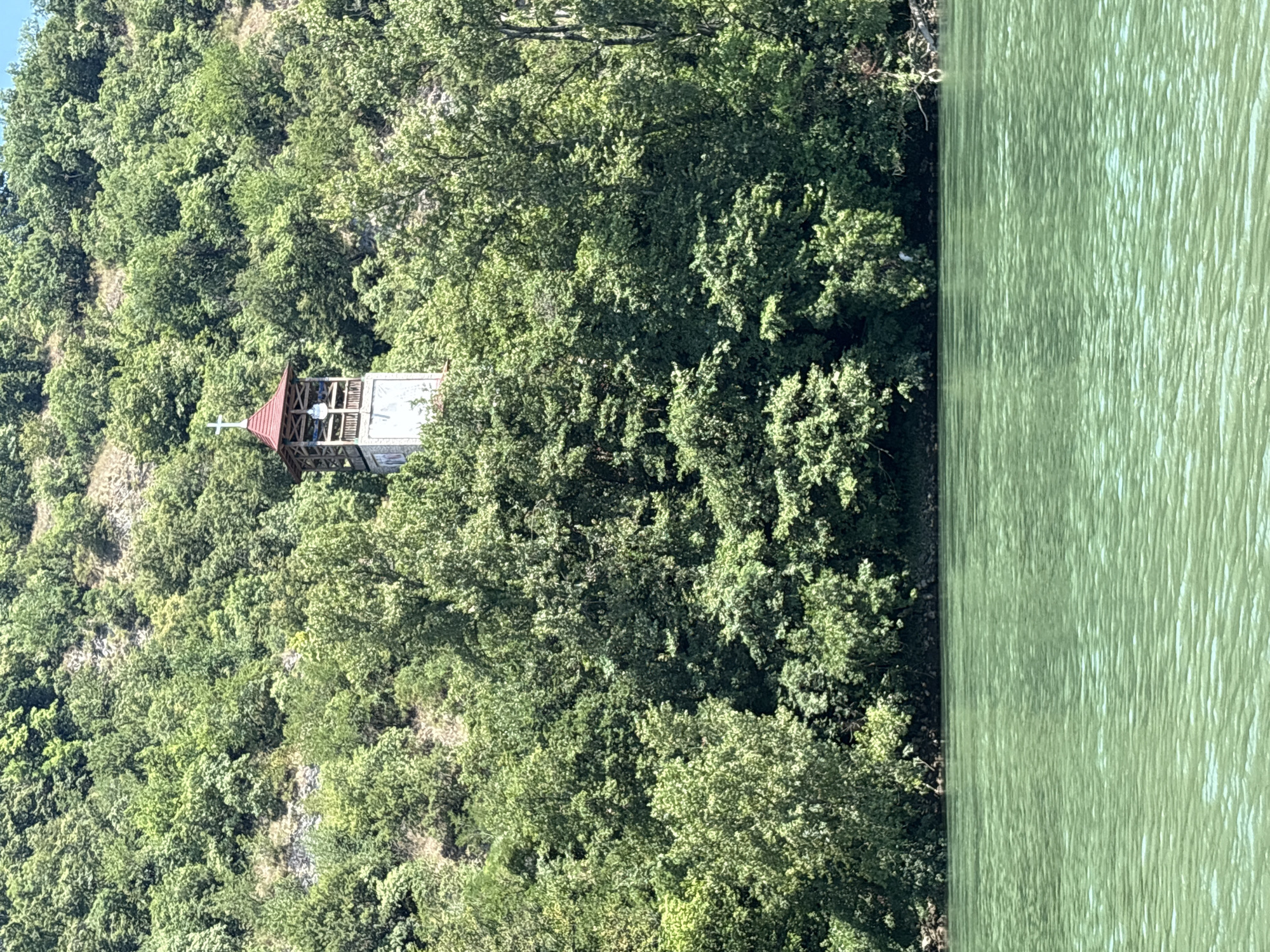 The image depicts a bell tower with a red roof and a cross, nestled amongst lush green trees on a steep hillside. The bell tower is a prominent structure, appearing to be made of stone and wood, with open arches revealing a glimpse of what might be a bell or a statue within. Below the hillside, a calm, greenish body of water stretches across the foreground, reflecting the light and creating a textured surface. The scene suggests a serene natural environment, likely a location overlooking a river or lake, possibly in a rural or semi-rural setting. The specific location in Zvornik, Bosnia and Herzegovina, is not visually identifiable from the image alone, but the presence of a religious structure on a hillside overlooking water is consistent with many scenic locations in the region.