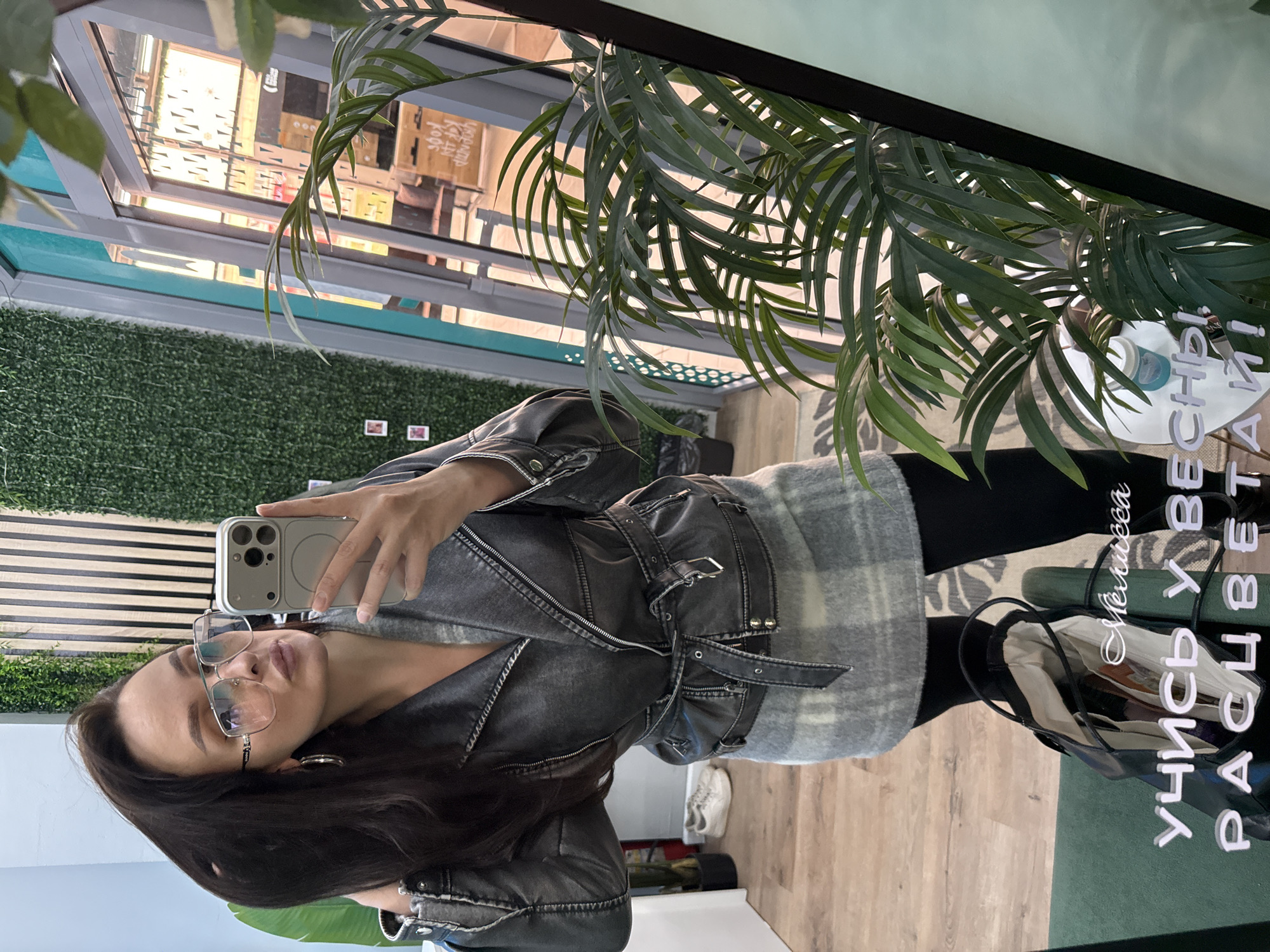 A young woman is taking a mirror selfie in what appears to be a shop. The environment is modern, with decorative greenery and a shop window reflecting external elements, potentially indicating an urban setting in Chaikovsky, Russia. The woman is wearing glasses, a black jacket with buckle details, and a plaid scarf. She holds a phone in her hand, presumably to capture the image. The interior decor includes artificial plants and striped wall panels. Near the bottom of the frame, a bag is visible, along with flooring that seems to be wooden. Writing is reflected in the mirror, suggesting a Russian-language inscription.