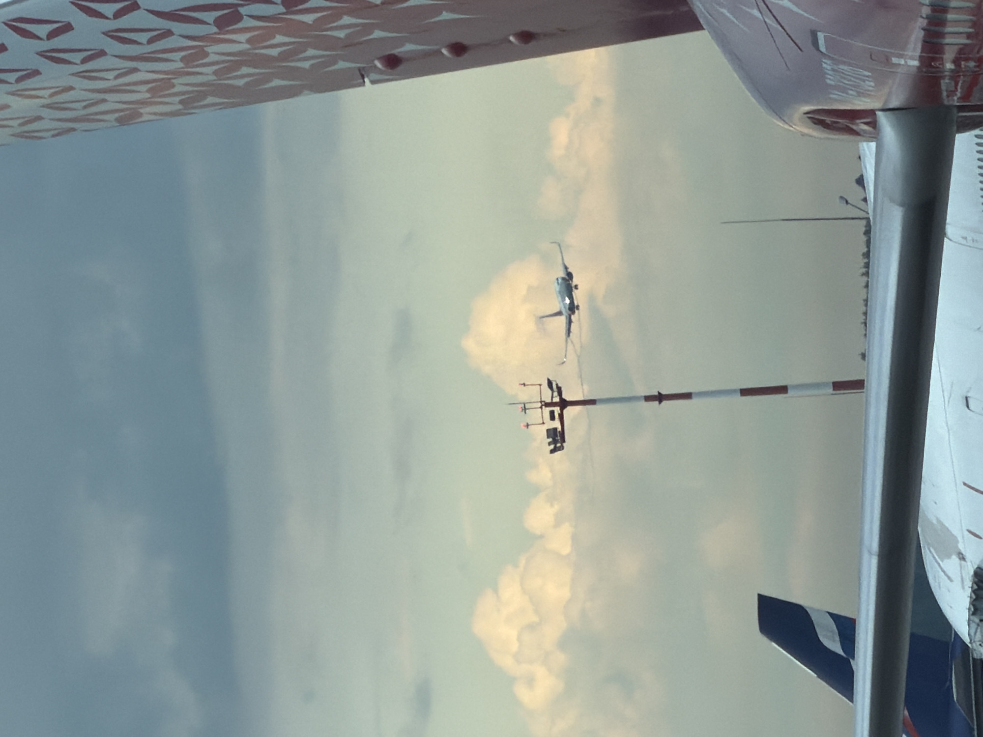 In Khimki, Russia, at an airport, a light blue airplane is captured in mid-air against a cloudy sky. In the foreground, the tail fin of a red and white patterned airplane is visible on the right side of the image, and a portion of a white airplane wing extends across the lower center. To the left, the tail of a blue airplane with a white stripe is partially visible. A tall, red and white striped pole with lights and equipment stands in the middle ground, and a smaller pole with a light is visible behind it. The overall scene suggests an active airport environment with aircraft in various stages of operation.