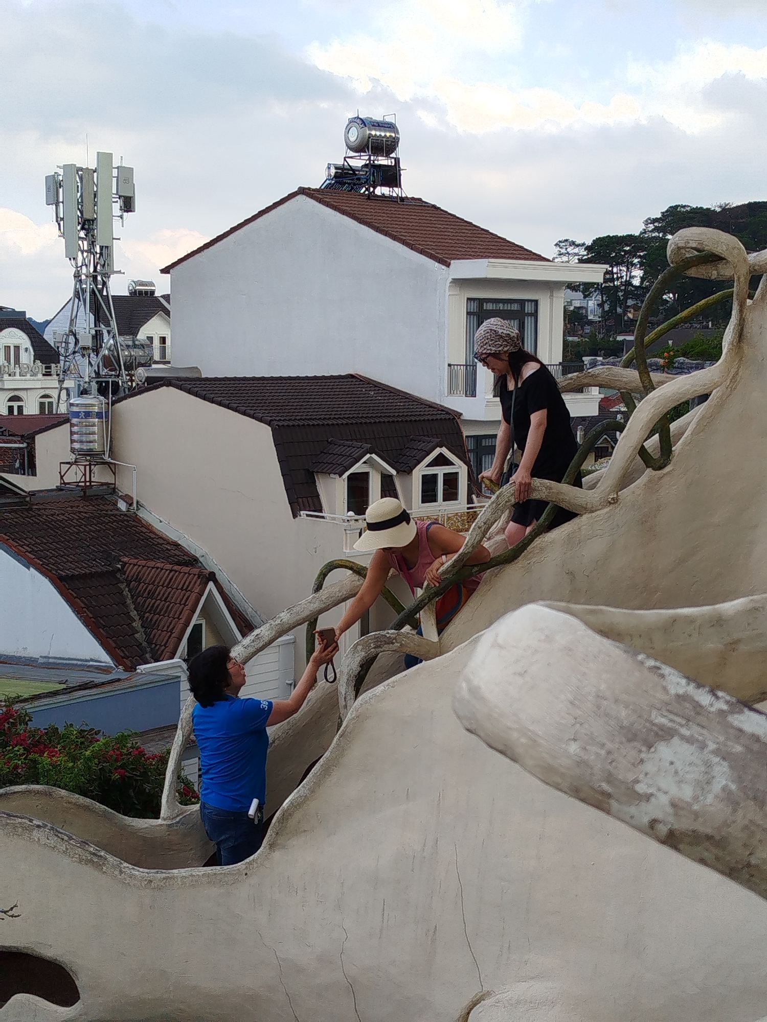 The image depicts a scene at the Crazy House in Da Lat, Vietnam. In the foreground, a person in a blue shirt is holding up a mobile phone, seemingly taking a picture of a woman wearing a sun hat and a pink top. She is climbing on the surreal, tree-like architecture of the Crazy House. Behind her, another woman in a dark dress and patterned head covering is also climbing on the structure. The Crazy House architecture is a prominent feature with its organic, cave-like design. In the background, buildings with traditional Vietnamese architecture, including tiled roofs and white walls, can be seen. A telecommunications tower is visible atop one of the buildings. The sky is overcast.