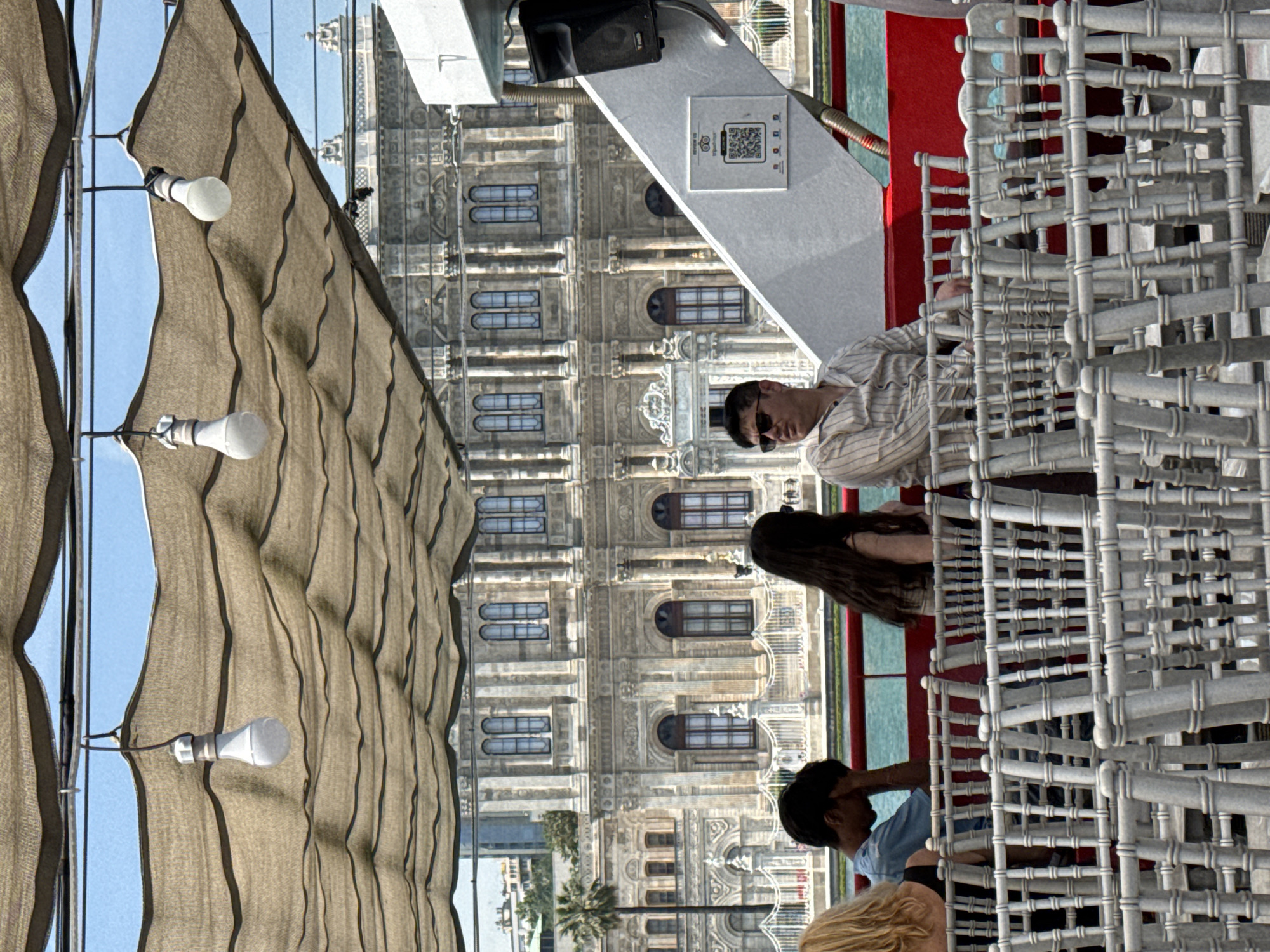The image captures a vibrant daytime scene from what appears to be a boat or ferry, offering a view of a grand architectural landmark. In the foreground, a brown, pleated canvas canopy provides shade, with several white LED lightbulbs hanging from its structure.
On the deck, a man in sunglasses and a light-colored striped shirt is seated, observing the view. Behind him, a woman with long dark hair is partially visible, while further back, another person in a blue shirt interacts with a blonde-haired individual. Stacks of white, intricately designed lattice chairs occupy a significant portion of the deck. A white counter or table is also visible, featuring a black speaker and a white rectangular sign displaying a QR code and the text "EL BAZAAR," alongside various small icons.
Across a body of blue-green water, a magnificent, multi-story stone building dominates the background. Its elaborate facade is adorned with numerous arched and rectangular windows, balconies, columns, and intricate carvings, characteristic of ornate Ottoman or European-influenced architecture, strongly resembling Dolmabahçe Palace. Palm trees can be seen near the building, indicating a warm climate.
The activity suggests people are enjoying a leisurely cruise or seated at a waterside establishment, taking in the picturesque views of the historic architecture under a clear blue sky. The scene is bright, indicating good weather during the day. The location, given the architectural style and water setting, is consistent with the Bosphorus strait in Beşiktaş, Turkey.