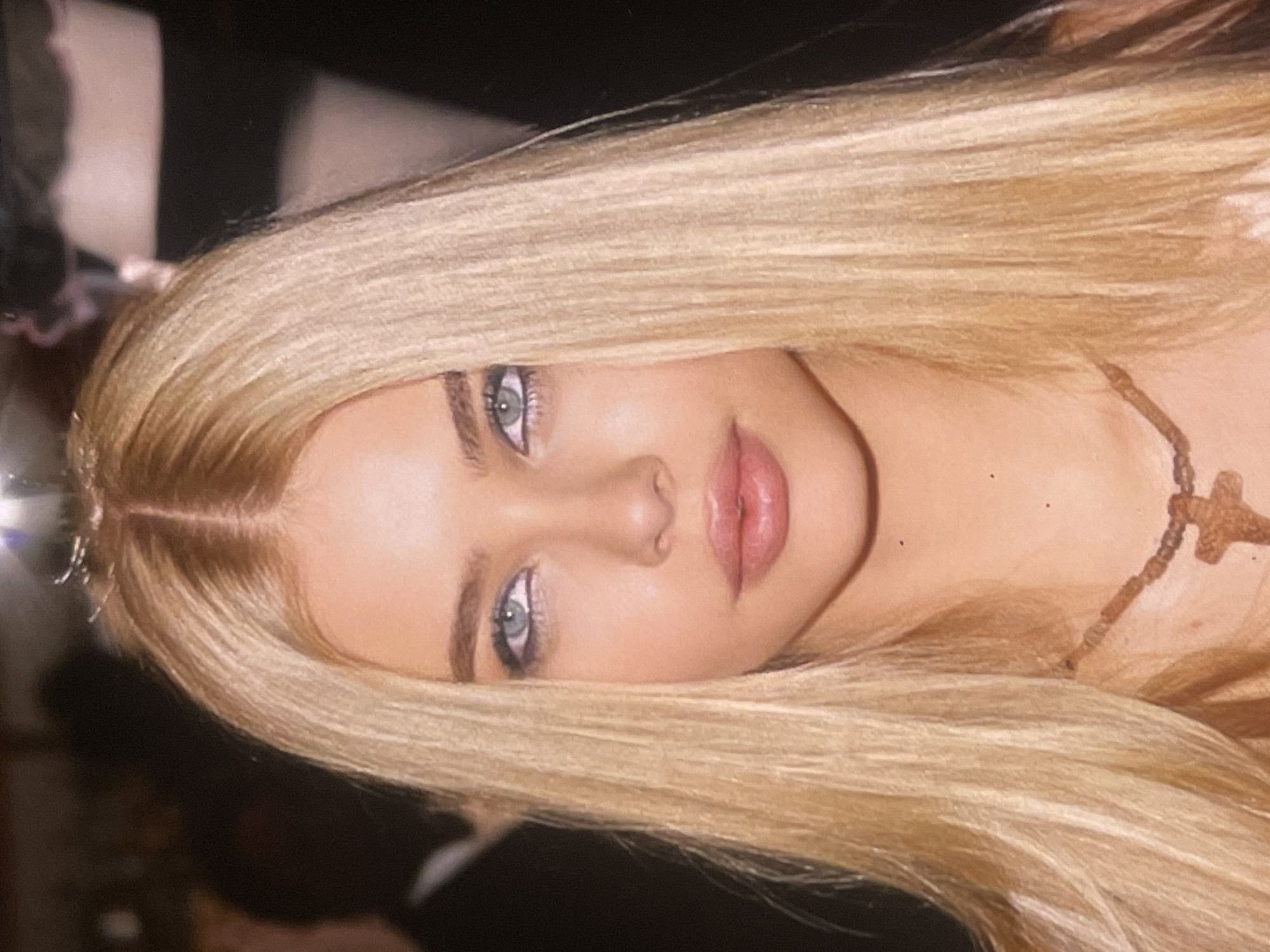 A close-up portrait shows a person, likely female, with fair skin and prominent features. Her long, straight, light blonde hair, parted in the middle, frames her face. She has striking light blue or grey eyes, enhanced with dark eyeliner and mascara. Her lips are full and appear to have a natural pink color with a glossy finish. Around her neck, a delicate necklace featuring a light brown, possibly wooden, cross-shaped pendant is visible. The background is dark and out of focus, suggesting an indoor setting or low light conditions, and it contains indistinct blurry shapes. The person is looking directly at the camera with a neutral expression, posing for the photograph. The image is rotated ninety degrees clockwise. There are no visible location cues or text within the frame.