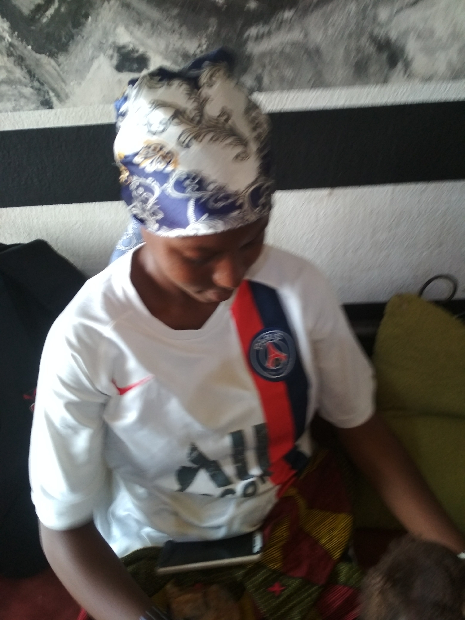 A person with dark skin is seated indoors. They are wearing a white t-shirt with a red and blue stripe down the front, a logo on the chest, and what appears to be a headscarf with a white, blue, and gold pattern. The person is looking down, holding a phone, and sitting in a chair or on a sofa covered with patterned fabric. Behind them, there's a wall with a horizontal black stripe over a white one and what looks like an abstract painting above. A dark-colored bag is to the left, and an olive-green cushion is to the right. The location and city are unknown.