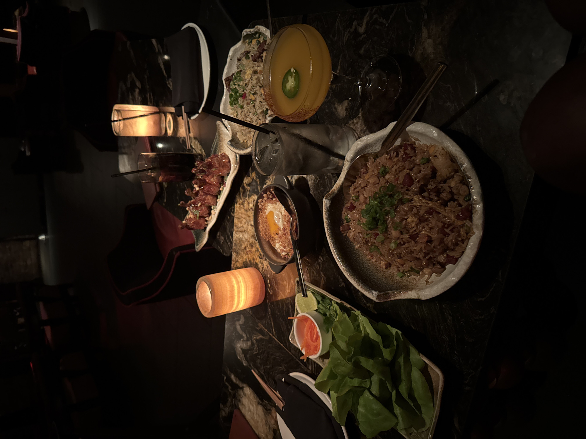 This image captures a dimly lit, intimate dining scene at what appears to be a restaurant or bar, likely at night. A dark, marbled table is laden with an array of Asian-inspired dishes and drinks.
Prominently featured are two large bowls of fried rice, one topped with chopped green onions. Beside them, a platter holds what looks like crispy fried chicken or shrimp, garnished with crushed peanuts. Another dish contains fresh green lettuce leaves accompanied by a small bowl of shredded carrots and lime wedges, suggesting a component for wraps. A smaller bowl displays a fried egg, possibly intended as a topping for a dish or as an appetizer.
Beverages include a martini glass filled with a yellowish cocktail, garnished with a slice of jalapeño, and a glass of water with ice and a straw. Another empty glass is partially visible on the table.
The scene is illuminated by two amber-glowing table lamps, casting a warm, inviting glow and reflections on the dark table surface. Chopsticks and other cutlery are also present, indicating a meal in progress. No people are visible, but the setup suggests a social dining experience for at least two individuals. There is no discernible text within the image. The overall ambiance is relaxed and atmospheric.