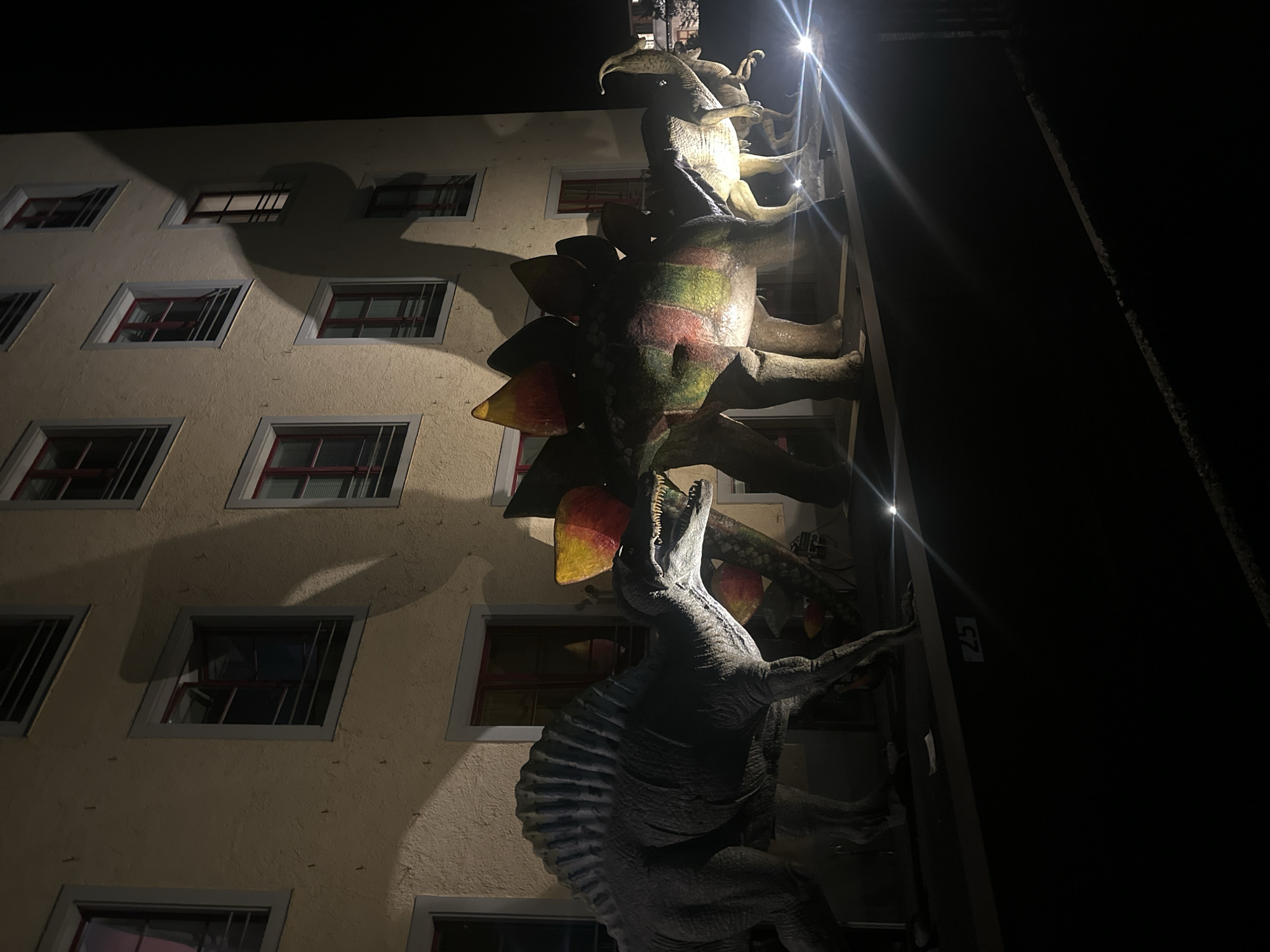 Captured at night in Sack, Switzerland, the image depicts life-sized dinosaur sculptures affixed to the exterior wall of a building. The dinosaurs, including a Stegosaurus and Dilophosaurus, are illuminated by nearby spotlights. The building, multiple stories tall, has windows with red frames and bars. The sculptures are attached to the building's facade via a dark-colored structure. The background is dark, showing little detail beyond the adjacent building's wall.