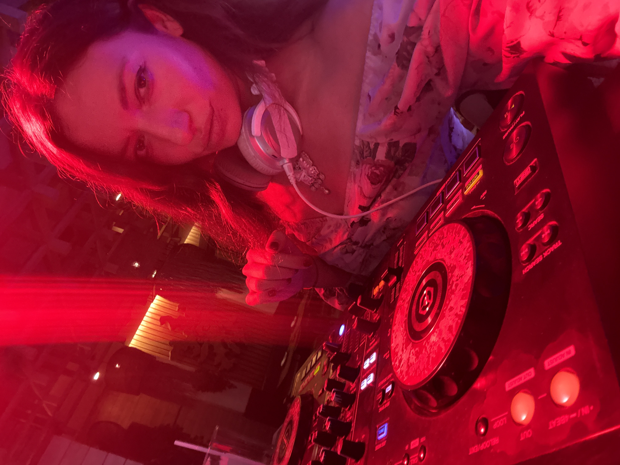 A woman, illuminated by red lighting, poses for a selfie in front of a DJ console. She has long, dark hair and is wearing headphones around her neck, which are adorned with a small, silver charm. The DJ console, with its various buttons, knobs, and illuminated displays, is the prominent object in the foreground. The background is dimly lit, with hints of wooden paneling and what appears to be a bar area, suggesting a nightclub or entertainment venue. The overall atmosphere is energetic, implied by the lighting and the subject's confident pose. The location is likely a club in Ko Samui, Thailand, given the context of the image.