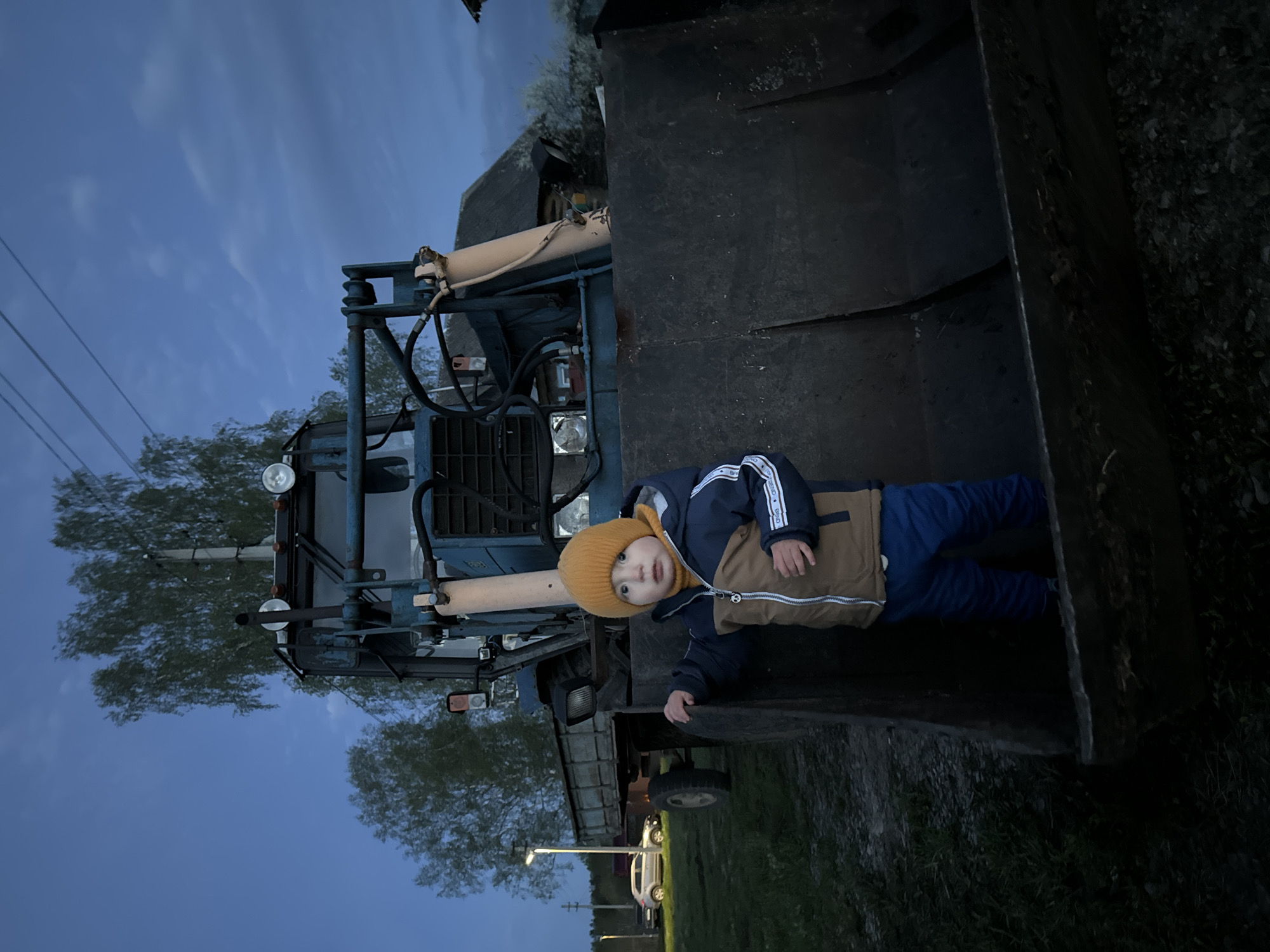 A young child wearing a yellow beanie, brown and navy jacket with white stripes on the sleeves, and blue pants is positioned in the bucket of a blue tractor. The tractor is parked outdoors in an area with trees and a utility pole. The sky is visible in the background, suggesting it is dusk or dawn. The location is Красный Маяк, Russia.