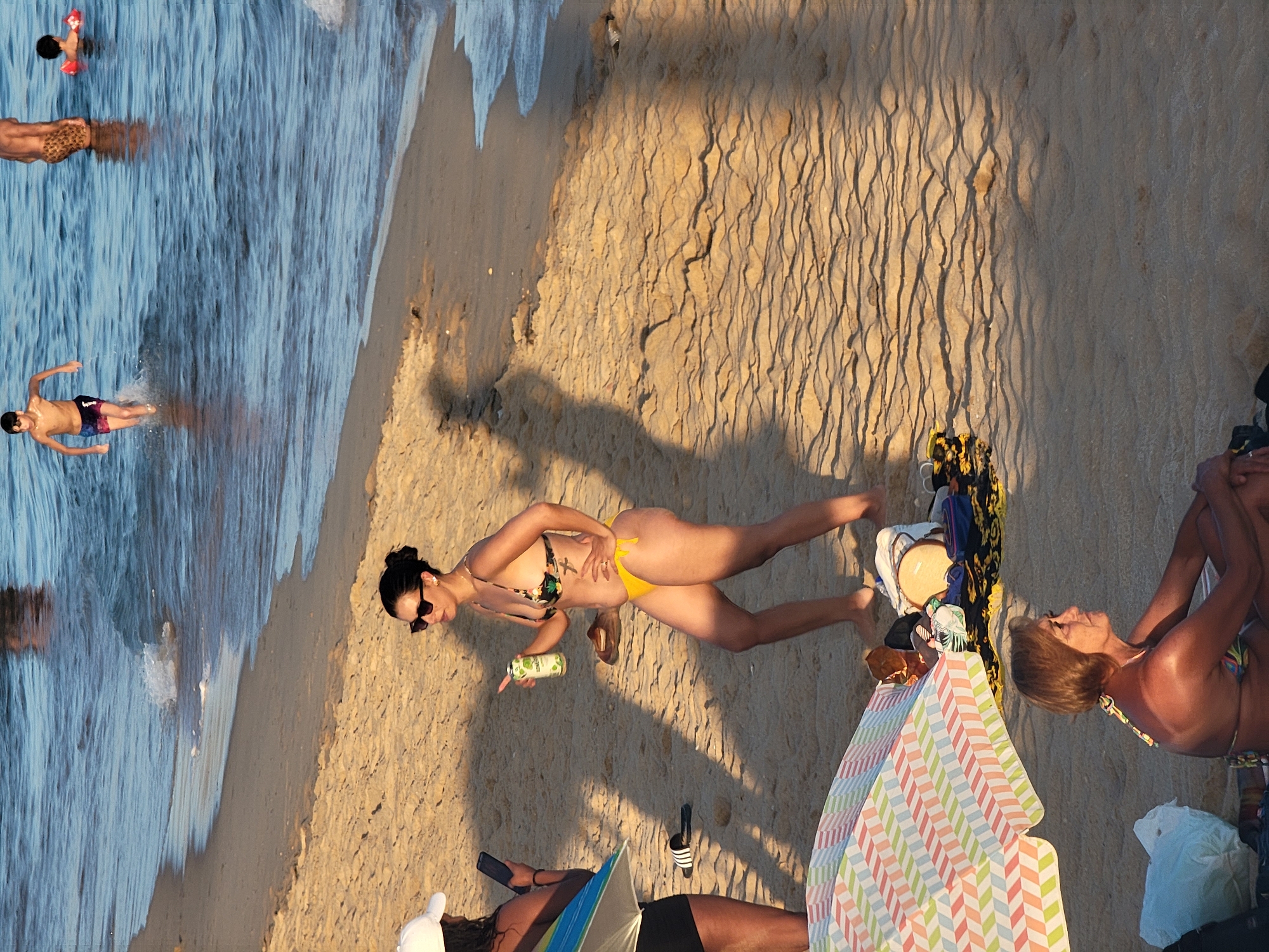 The image captures a sunny day at Quarteira beach in Portugal. In the foreground, an older woman with short brown hair sits on the sand, wearing a bikini and looking to the right. To her left, a colorful beach umbrella shades a pile of beach items, including a towel and bags. Further up the beach, a woman in a yellow bikini and sunglasses holds a can of drink and stands with her hand on her hip, looking to the left. Next to her, another woman wearing a white cap and black shorts holds a phone. In the background, several people are visible in the shallow water, including two young boys running and playing. The sea has gentle waves, and the sand is golden and rippled. The scene is illuminated by natural sunlight, casting long shadows across the sand.