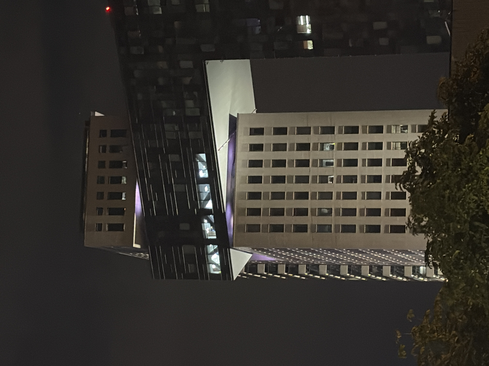 A nighttime, slightly angled, low-angle shot of a modern, multi-story building in Moscow, Russia. The building has a complex, layered design. The bottom portion of the visible building is a tall, rectangular tower with numerous evenly spaced windows. Above this, a large section of the building juts out, creating a horizontal overhang. This overhang has a dark, ribbed facade and many windows. A triangular white structure is visible beneath the overhang, connecting it to the lower tower. At the very top of the visible structure is a smaller, rectangular section with fewer, larger windows. The building is illuminated, with lights visible in many windows. The sky is dark and overcast, with only a faint red light visible on a distant part of a building to the right. Green foliage is visible in the foreground at the bottom of the image.