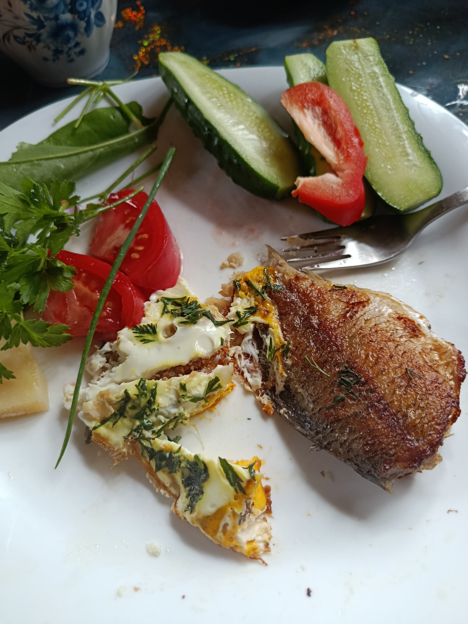 A close-up, overhead shot shows a white plate with a meal. The meal consists of a piece of fried fish, a portion of what appears to be scrambled eggs with herbs, sliced tomatoes, sliced cucumber, a slice of red bell pepper, and a small piece of cheese. A fork rests on the plate. The background is blurred, but appears to be a dark surface, possibly a table, with some decorative elements. The image is taken indoors in the city of Vereya, Russia.