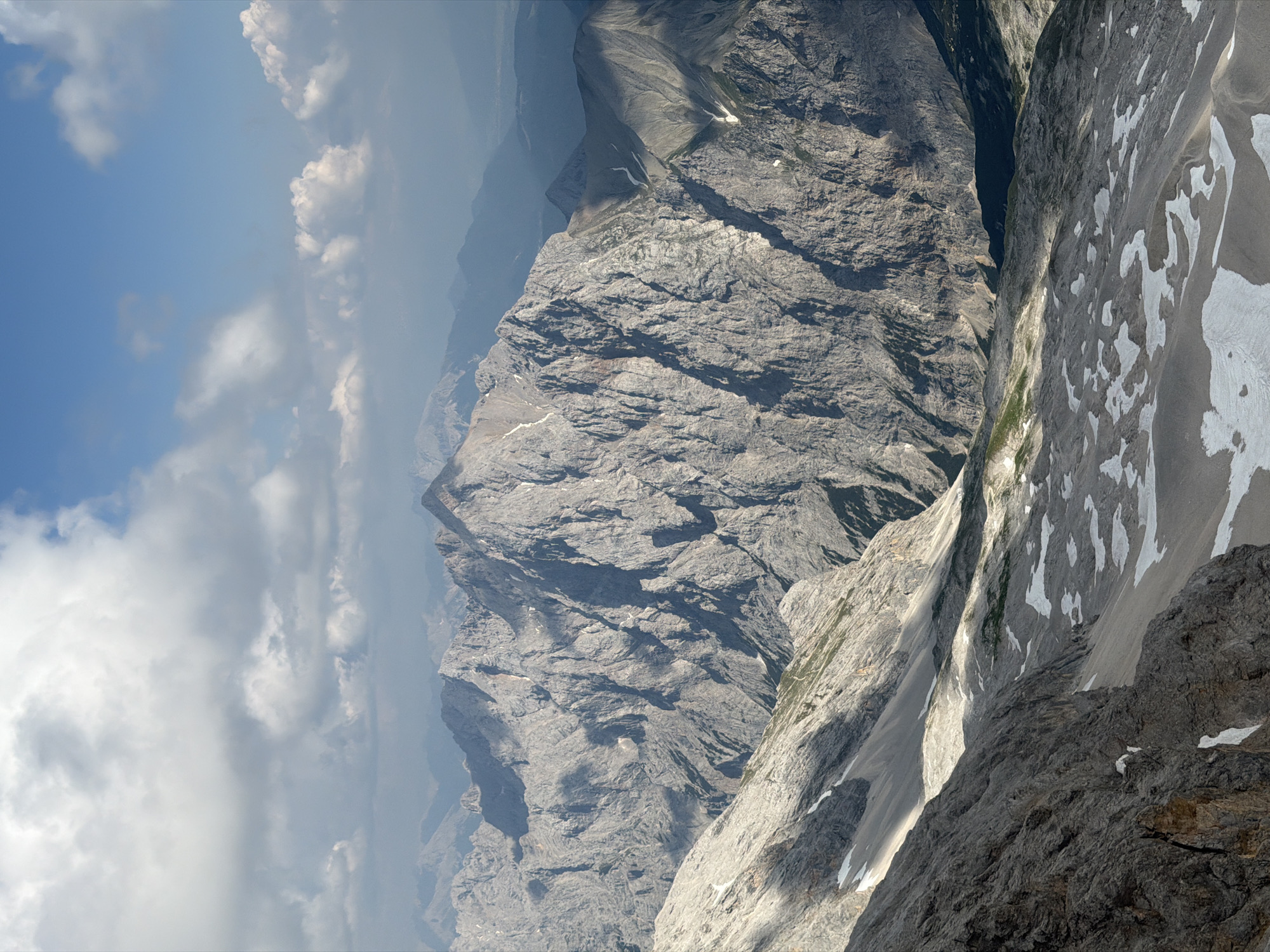 This media file captures a stunning, panoramic view of a majestic mountain range, likely in the Garmisch-Partenkirchen region of Germany. The scene is dominated by rugged, grey rock formations, weathered and carved by time and the elements. Patches of snow cling to the higher slopes, stark white against the dark stone, indicating a cool or alpine environment. The terrain is steep and dramatic, with deep ravines and sharp peaks.
The sky above is a blend of brilliant blue and soft, ethereal clouds, suggesting a partly cloudy day with good visibility. The sunlight casts dramatic shadows across the mountainsides, highlighting their texture and scale, and indicating it is likely midday or early afternoon. The overall impression is one of immense natural grandeur and isolation. There are no people or man-made objects visible in the frame, emphasizing the wild and untamed nature of the landscape. The image conveys a sense of awe and the vastness of nature.