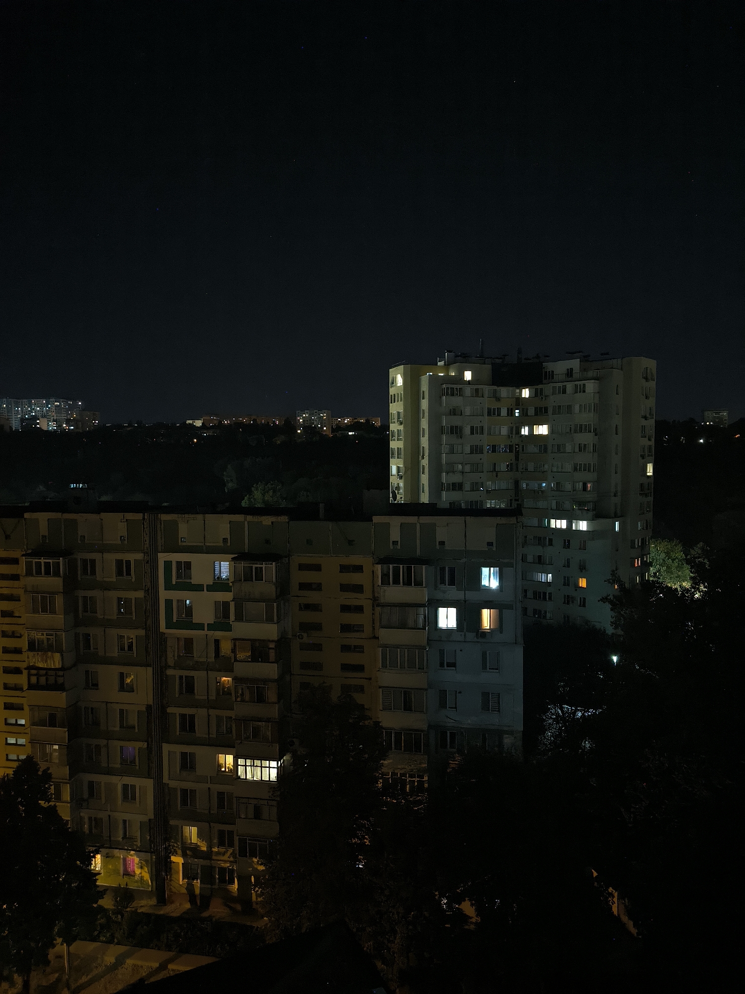 An aerial nighttime shot captures a dense urban landscape in Chișinău, Moldova, dominated by apartment buildings and the dark expanse of the sky. The foreground features a long, multi-story apartment building with numerous windows, some illuminated with warm, yellow light, suggesting occupancy. To its right and slightly behind it, a taller, cream-colored apartment building stands, also dotted with brightly lit windows. In the background, more buildings and distant city lights are faintly visible against the dark sky, interspersed with trees and vegetation that create deep shadows. A few faint stars are visible in the upper portion of the image. The overall scene conveys a sense of urban life continuing into the night, with pockets of light illuminating residential areas.