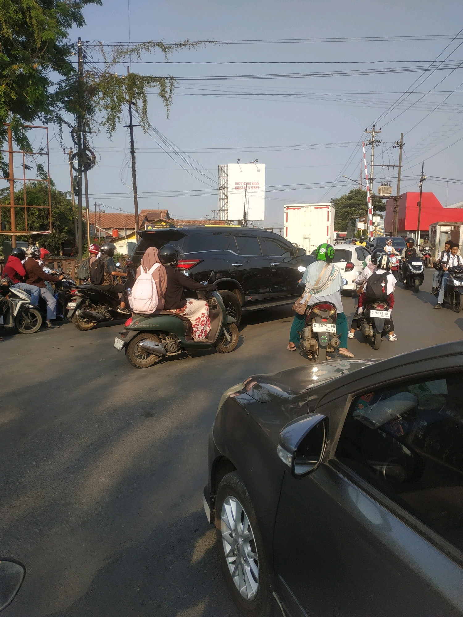 The image depicts a street scene in Tegal, Indonesia, likely during daylight, with a traffic jam. A dark SUV is prominent in the center, surrounded by numerous motorcycles. Several individuals are riding on these motorcycles, some wearing helmets, with varying attire. One motorcycle in the foreground is green, carrying two people, one with a pink backpack. Another motorcycle behind it has two riders, the rear passenger wearing a headscarf and carrying a pink backpack. Further back, a person in a light blue top and green helmet is on a motorcycle. A white truck is visible in the background, along with buildings and power lines. The road surface is asphalt, and sunlight casts shadows. There is a railway crossing barrier in the middle ground. The overall impression is one of congested urban traffic.