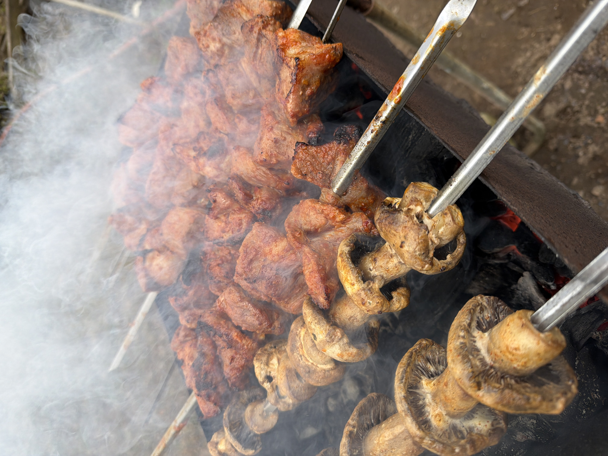 The image shows skewers of meat and mushrooms grilling over hot coals in what appears to be a barbecue setup outdoors. Dense white smoke partially obscures the meat skewers to the left, which are loaded with browned, cubed pieces. The mushroom skewers to the right are closer to the camera and show large, whole mushrooms threaded onto metal skewers. A metal grilling rack is visible beneath the skewers and above the coals. The image is taken from above and at an angle, looking down on the food and the grill. The location Бронница, Russia is not discernible from the image itself.
