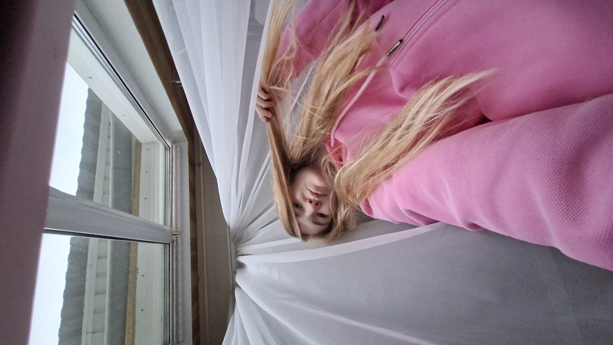 The media file shows a young woman with long blonde hair, wearing a pink hooded sweatshirt. She is positioned in what appears to be a room, with a window visible on the left and sheer white curtains on the right. The image is taken from a low angle, with the woman's head and upper body filling most of the frame, and her hair cascading down. Her face is turned towards the camera, and she has a neutral expression. The window shows an overcast sky and the exterior of a building with a textured facade. The overall lighting suggests it is daytime, but the overcast conditions might indicate a cloudy or possibly snowy day in Большой Букор, Russia. There are no visible text elements in the image.