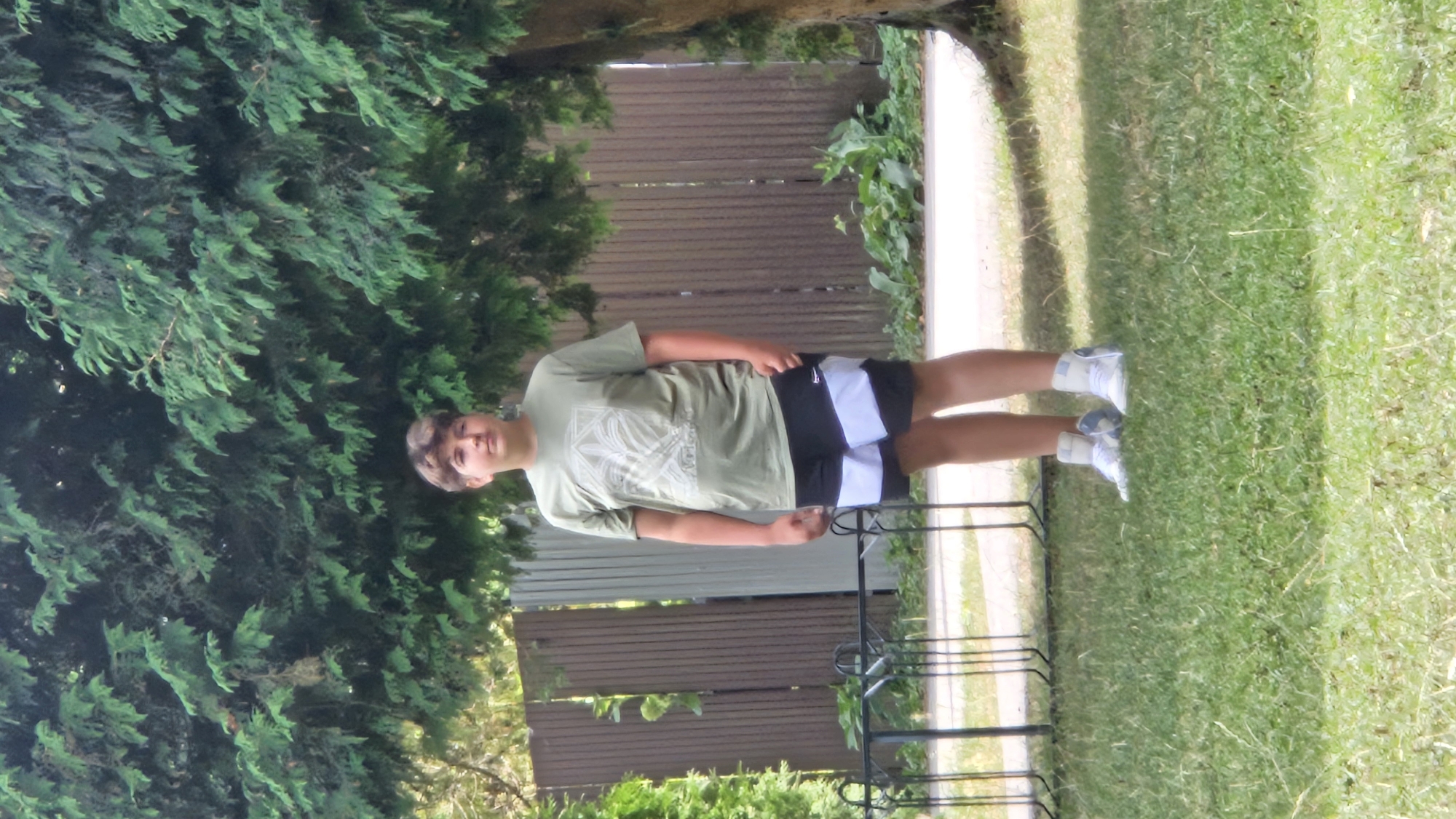 A young male, appearing to be in his early teens, stands in a grassy area with a large tree and a wooden fence behind him. He is wearing a light olive green t-shirt with a graphic design on the front, and black shorts with white horizontal stripes near the bottom. He is also wearing white high-top sneakers. The background features lush green foliage and a brown wooden fence. The scene appears to be outdoors, possibly in a residential area or a park. The location in the city of Prešov, Slovakia is not determinable from the image.
