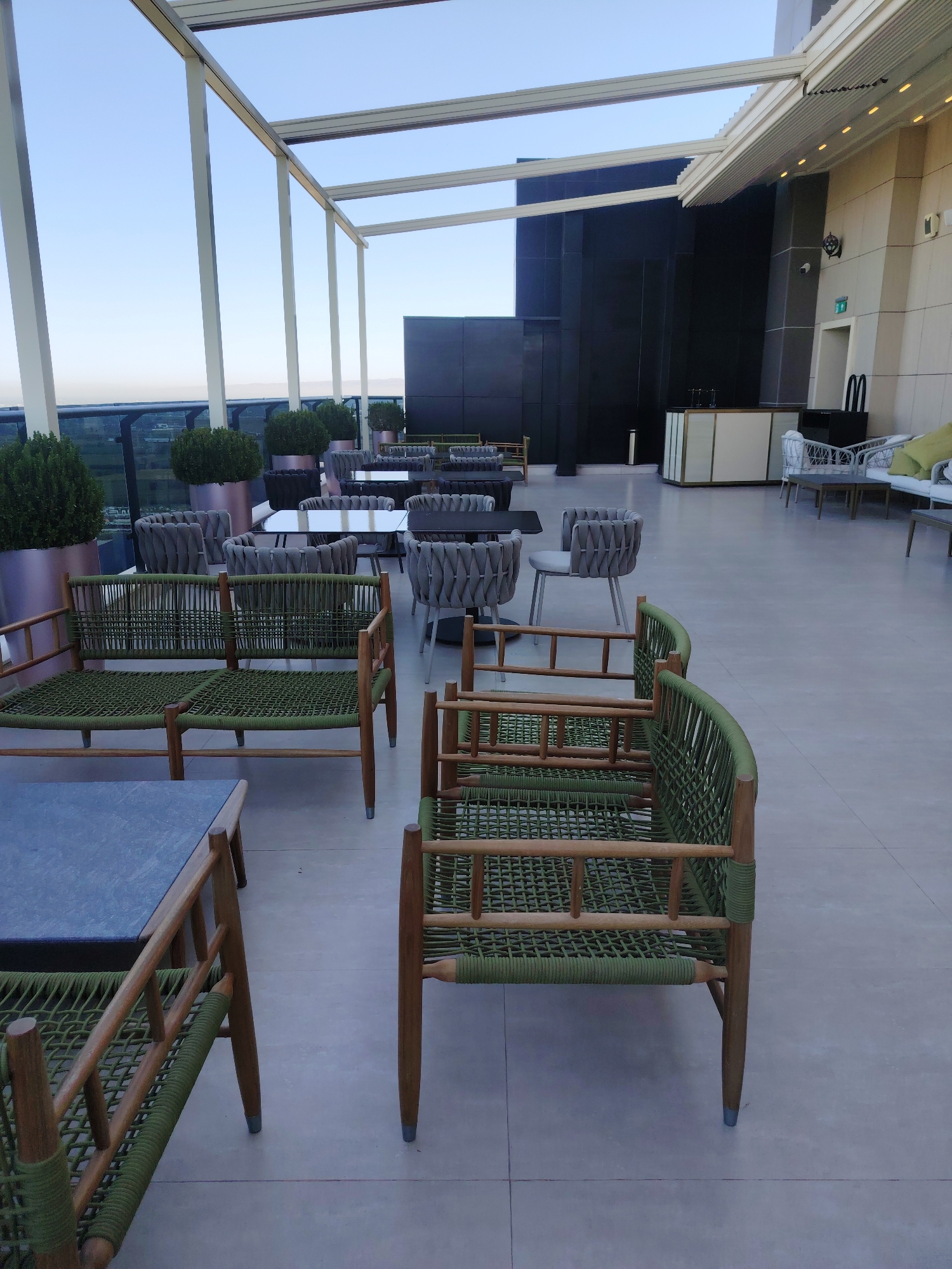 The image depicts a rooftop seating area in the city of Konigil, Uzbekistan. The scene is an outdoor patio furnished with various seating arrangements and tables, suggesting a cafe or restaurant setting. The foreground features a low wooden bench with green woven seating and backrests, alongside a dark-topped table. Further into the image, more seating is visible, including grey upholstered chairs with intricate woven backs, arranged around glass-topped tables. In the background, a modern building with dark facade panels rises into the sky, and a white outdoor canopy structure extends over the seating area. The sky is clear and pale blue, indicating daytime. There are no people visible in the image. The overall atmosphere is serene and organized, with the furniture arranged for relaxation and dining.
