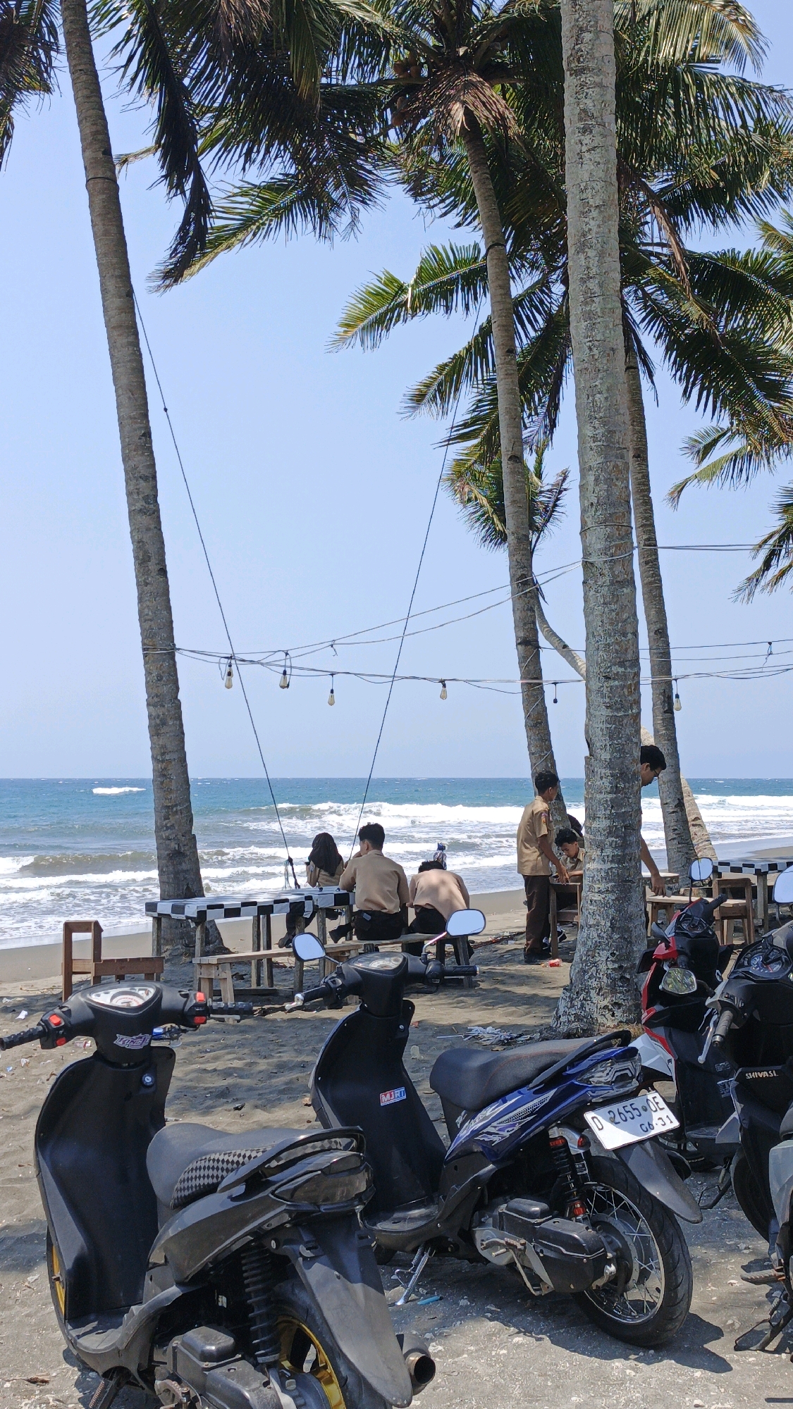 A group of people are gathered at a beachside cafe in Desa Batukaras, Indonesia, under the shade of palm trees. The ocean is visible in the background with moderate waves. Several motorbikes are parked in the foreground on the sand. Some individuals are seated at a table, while others appear to be standing or moving around. The lighting suggests it is daytime, with clear skies.