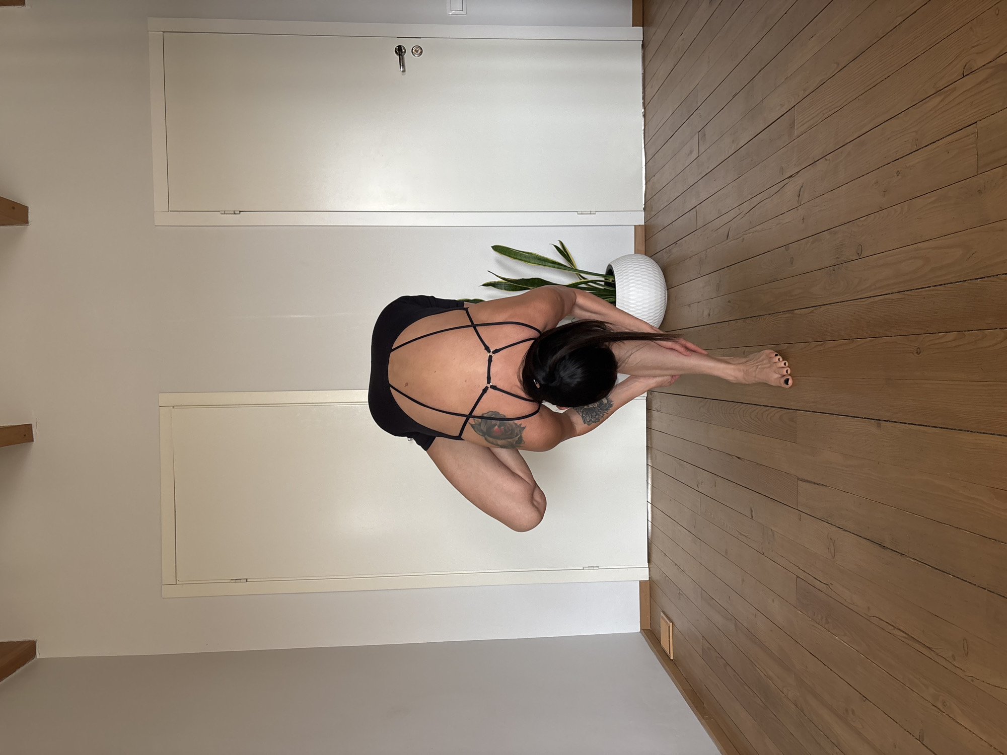 This high-angle image, captured in Chaikovsky, Russia, depicts an indoor scene, likely a room with a minimalist aesthetic. The setting features light, plain walls and a warm, light brown wooden plank floor.
The central subject is a person, positioned in the center-right of the frame, engaged in a yoga or stretching pose. They are bent forward with their head lowered towards their knees, which are flexed. Their arms appear to be resting on or lightly embracing their shins. The individual has dark hair, pulled back, and is wearing a black, possibly athletic, top or leotard with intricate, geometric strap detailing across the back. Visible tattoos include a floral design on the left shoulder/upper arm and another on the left forearm. One leg is extended forward, revealing a foot with dark nail polish on the toes.
To the right of the person, a green plant with tall, upright leaves, possibly a snake plant, is housed in a distinctive white, textured, spherical pot. The background features two white, flush doors or panels on the wall: a lower one with a silver door handle and keyhole, and a higher rectangular panel. In the upper-left corner, two small wooden elements protrude from the wall, possibly indicating shelves or structural features. The room is brightly lit, suggesting a daytime setting. No visible text is present in the image.