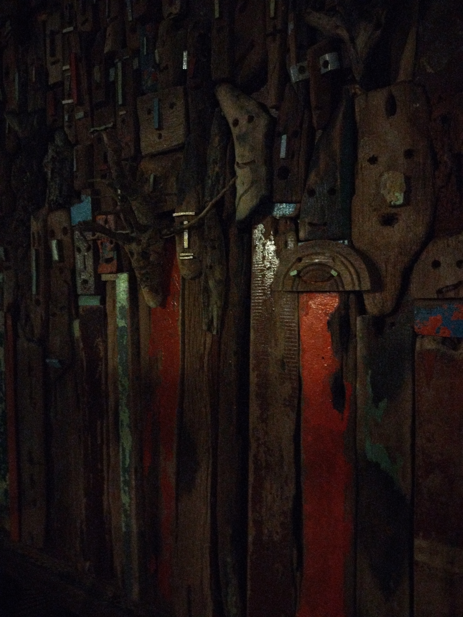 The image shows a wall constructed of various pieces of wood, many of which are carved into rudimentary face-like shapes. The wood is weathered and appears aged. There are splashes of paint, predominantly red, on some of the wooden planks. The lighting is dim, which creates shadows and obscures some details. The image was taken in ตำบลเกาะพะงัน, Thailand.