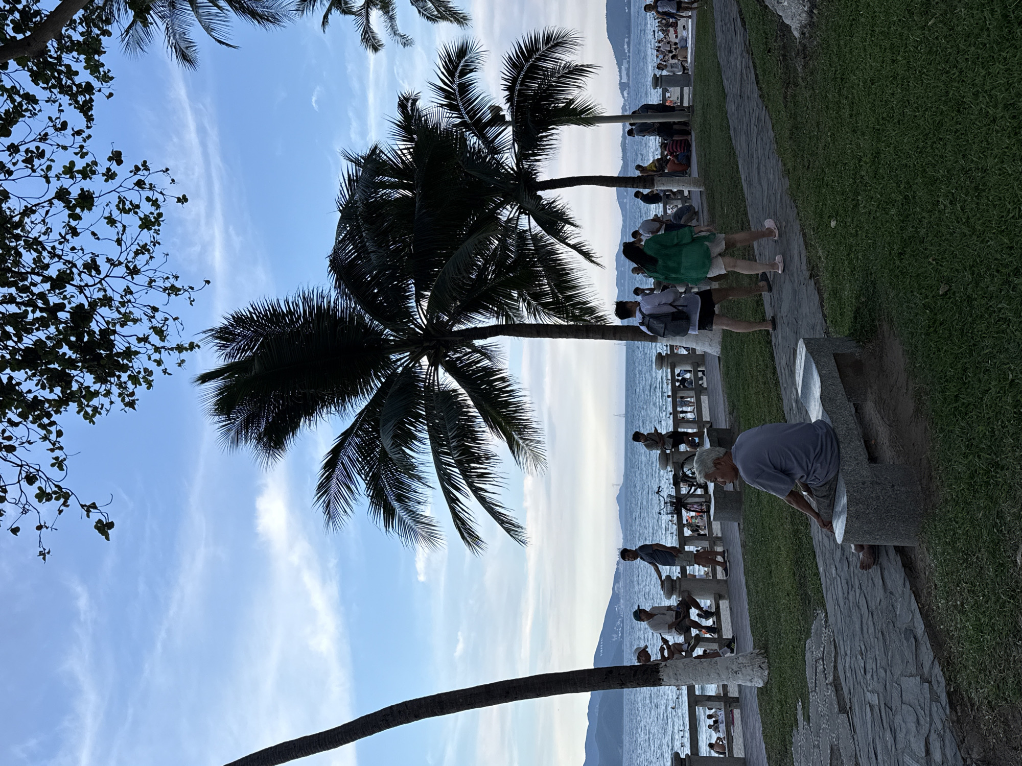 This image depicts a scenic coastal park in Nha Trang Ward, Vietnam. The scene is dominated by lush green grass and a stone pathway bordering the ocean. Several palm trees, their fronds silhouetted against a bright blue sky with wispy clouds, punctuate the landscape.
A number of people are present, scattered throughout the park. Some are strolling along the pathway, while others are seated on benches or near the water's edge, enjoying the view. In the foreground, an elderly man in a blue shirt is bent over a granite bench. Further down the path, a group of people, including a woman in a green top and shorts, are walking. Children can be seen on a lower level near the water. The presence of bicycles suggests a leisurely atmosphere.
The time of day appears to be late afternoon or early evening, given the soft light and the position of the sun, which is not directly visible but illuminates the sky. The weather seems pleasant, with a clear sky and calm waters. The distant mountains suggest a coastal city setting. The overall mood is relaxed and tranquil, with people enjoying a pleasant day outdoors. There is no visible text in the image.